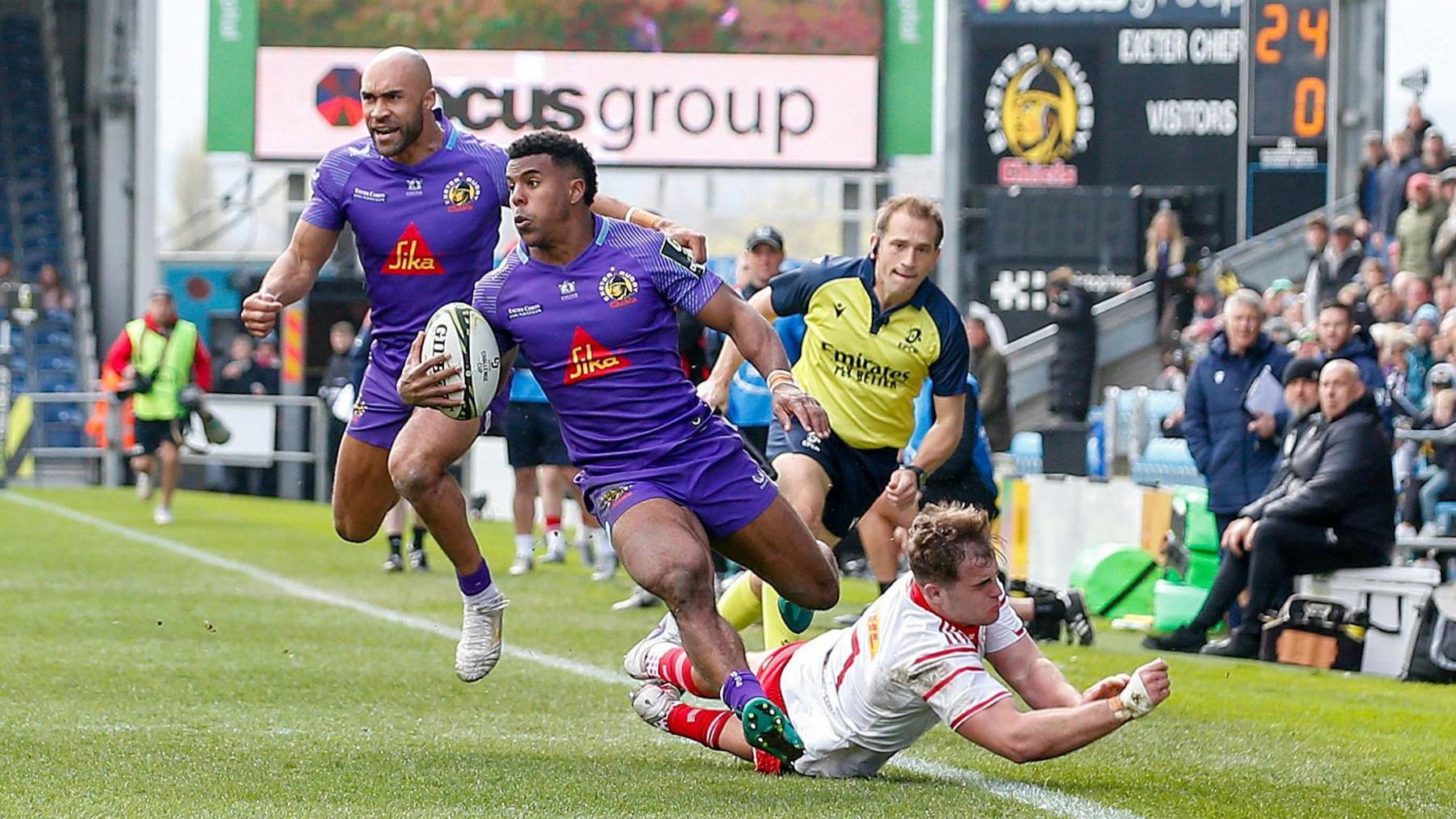 Immanuel Feyi-Waboso running around the diving Munster tackler on the left wing before scoring for Exeter with Olly Woodburn in the background as the linesman looks over to check if Feyi-Waboso's foot has gone into touch.