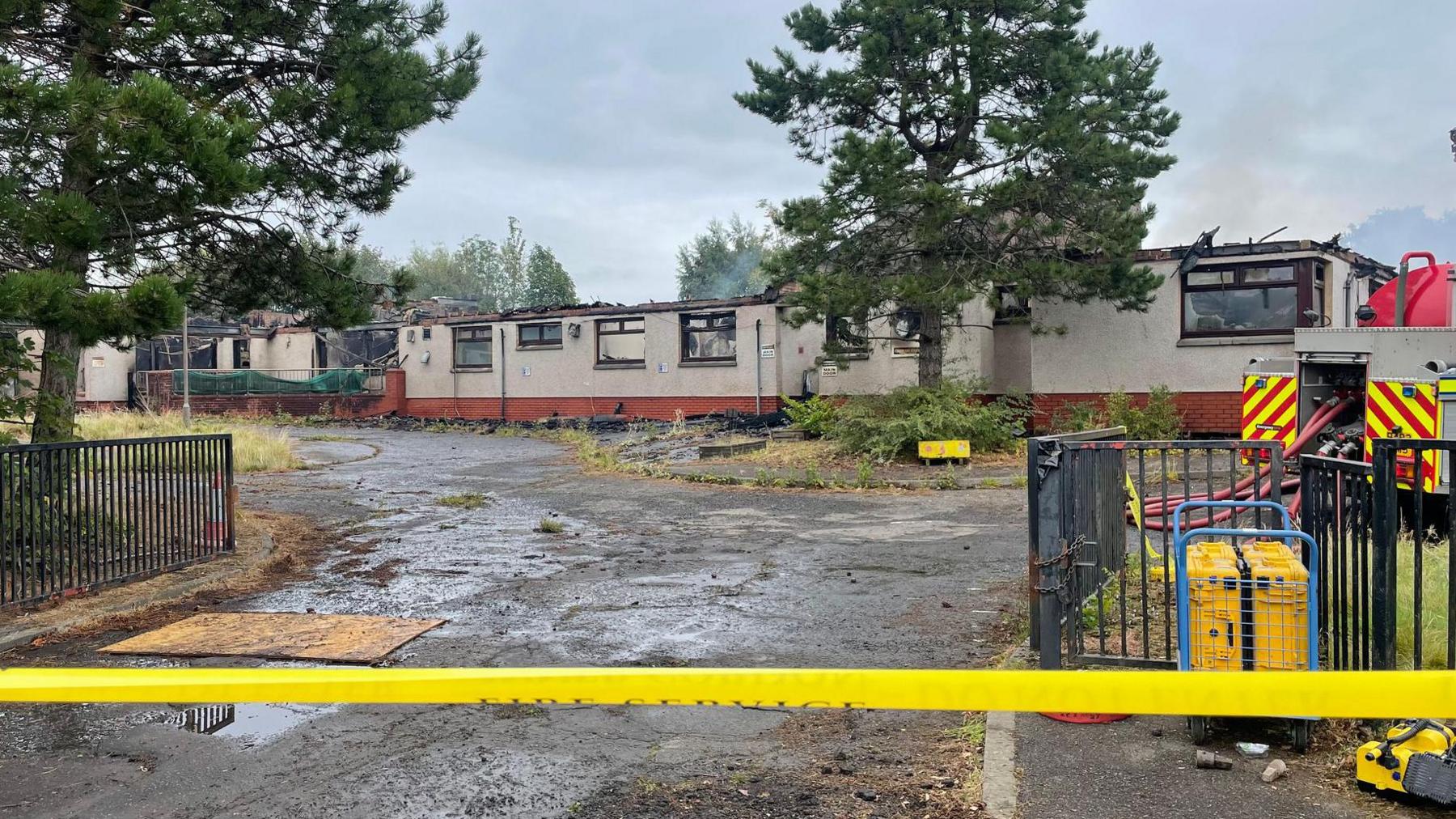 A burnt out one-storey building behind a yellow cordon. Trees are dotted around the building