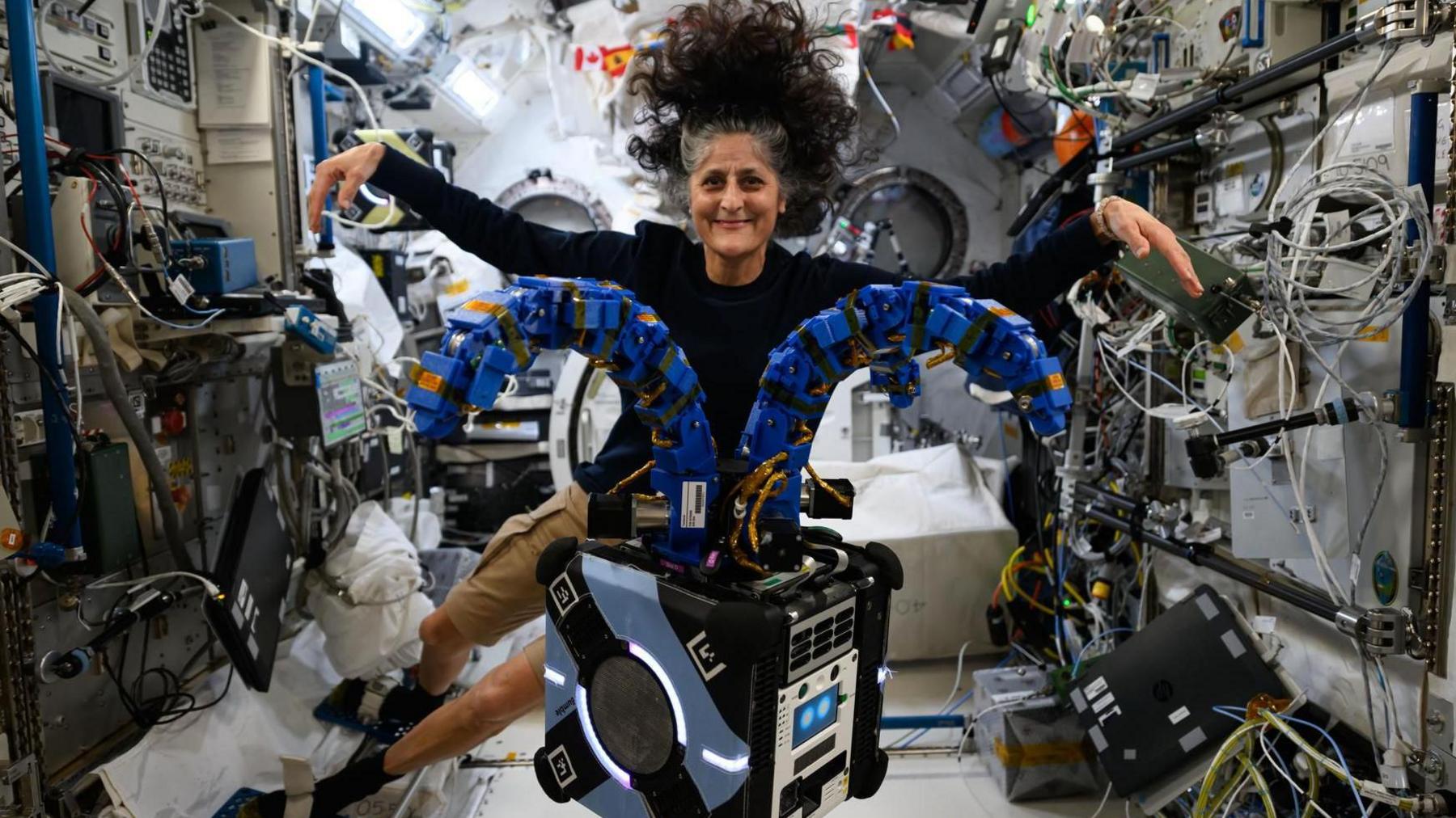 A smiling Suni Williams floating in the international space station, in a black long-sleeve top and beige shorts, hair wild and having a life of its own as it floats in zero gravity. Her arms are outstretched mimicking a box-like apparatus floating in front of her which has two thick arm-like blue appendages emerging from its top and spreading out. In the background the interior walls of the ISS are a messy riot of different coloured cables, keypads and monitors.