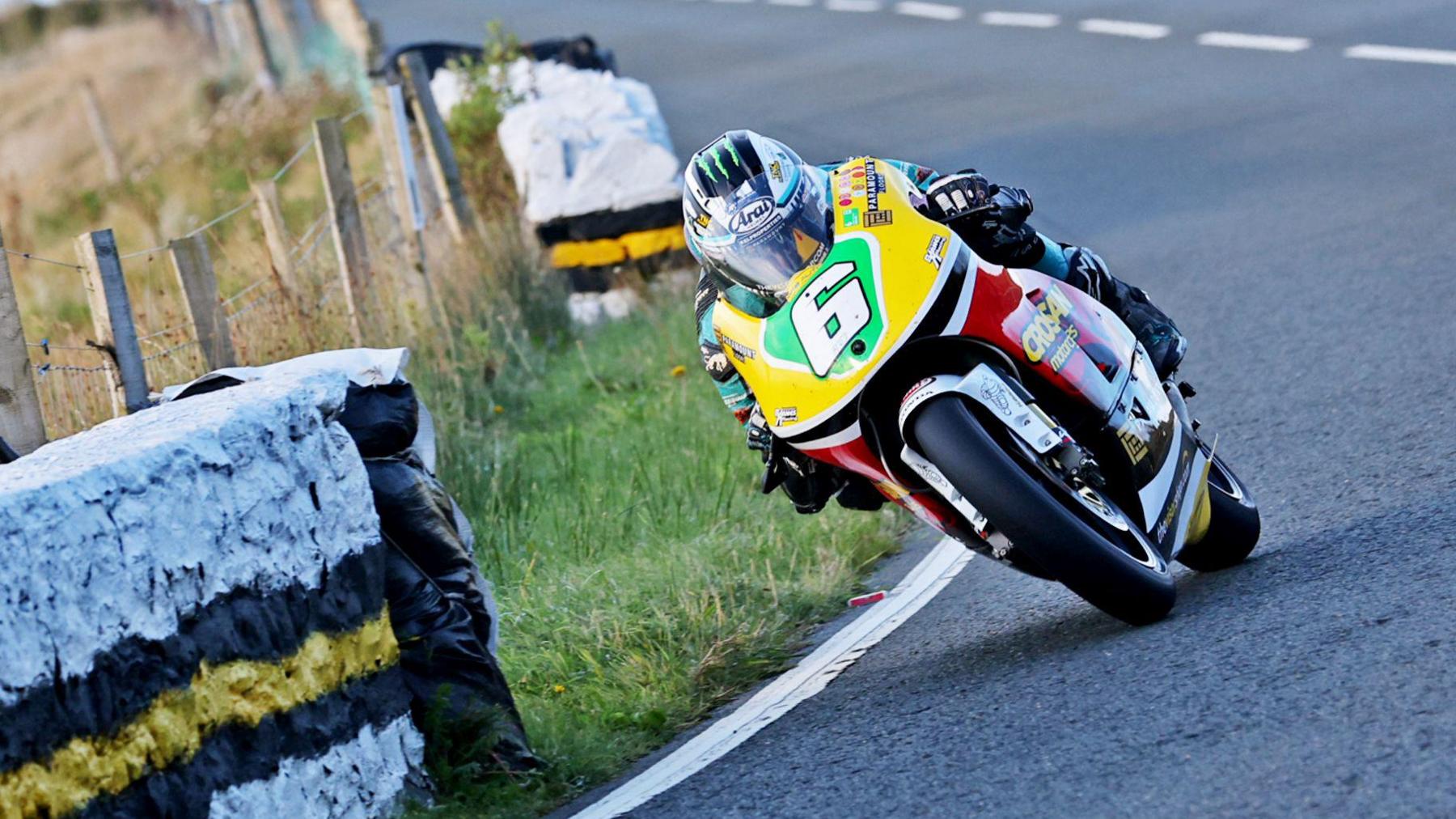 Michael Dunlop on the Lightweight machine, which is red and yellow and has the number six on a green plate on the front. He is leaning over as he passed a stone wall.