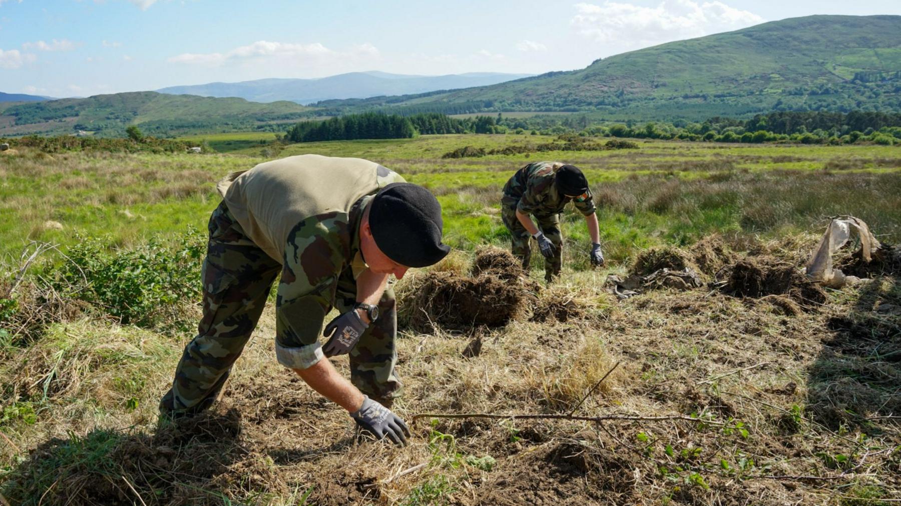 Two people wearing army clothing are digging in the ground with there hands. They are wearing gloves. In the background we can see a collection of hills and rugged countryside land. 