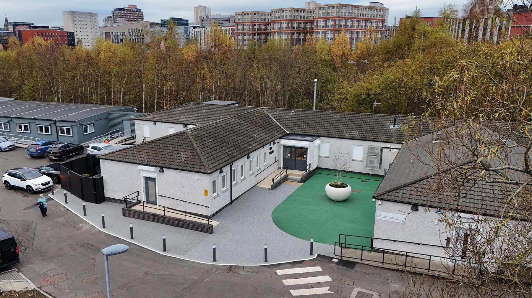 A drone image of the safe drugs consumption room in Glasgow. The building is white with a dark roof. It is in a U-shape with a green central courtyard and a large, white, plant pot in the middle. It is surrounded by grey paving. In the background, trees with green and orange leaves can be seen alongside several high rise buildings.