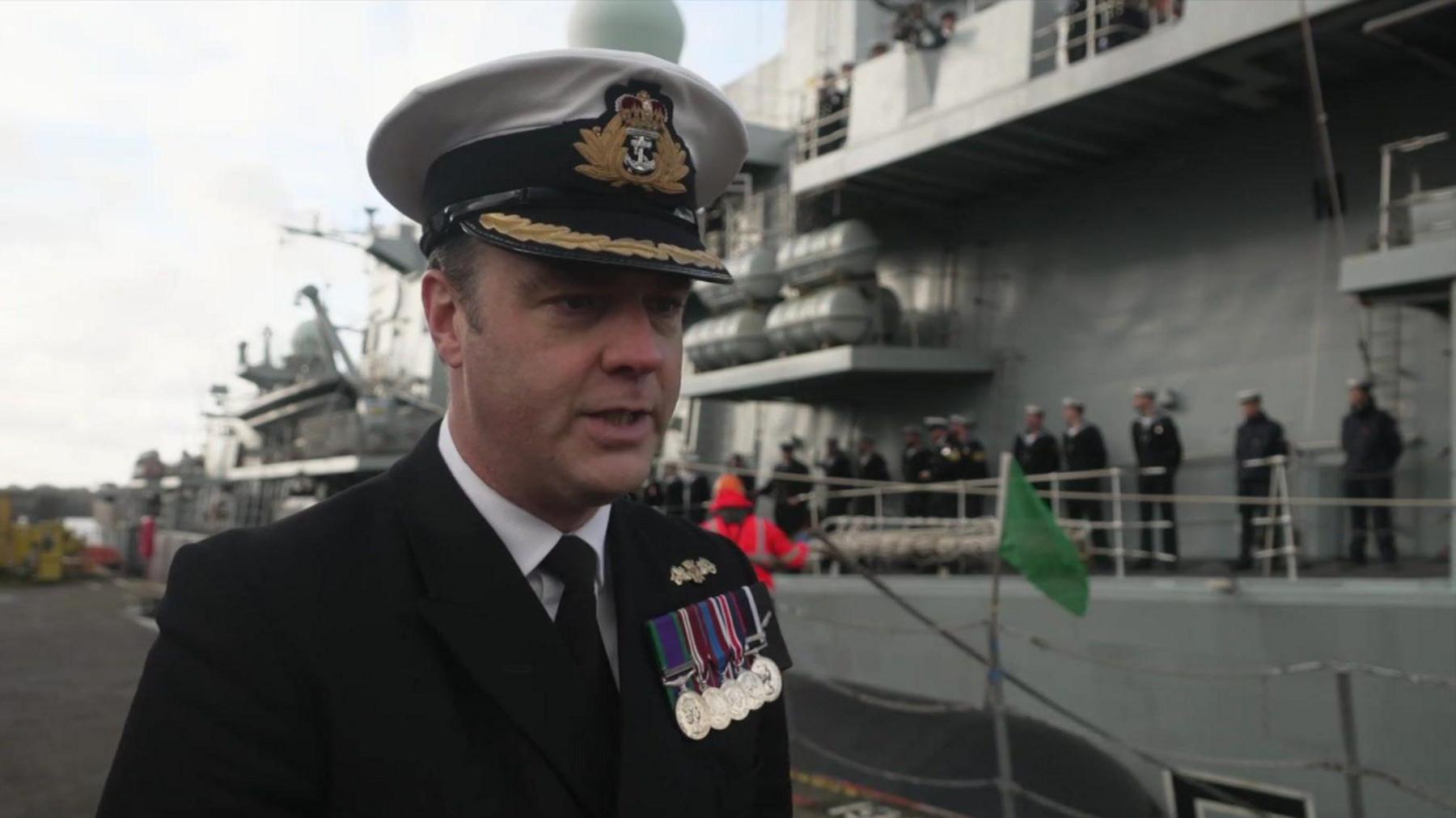 Ian Feasey looking to the right. He is standing on the dock in front of the frigate. The frigate is grey and there are sailors standing along the side of it.