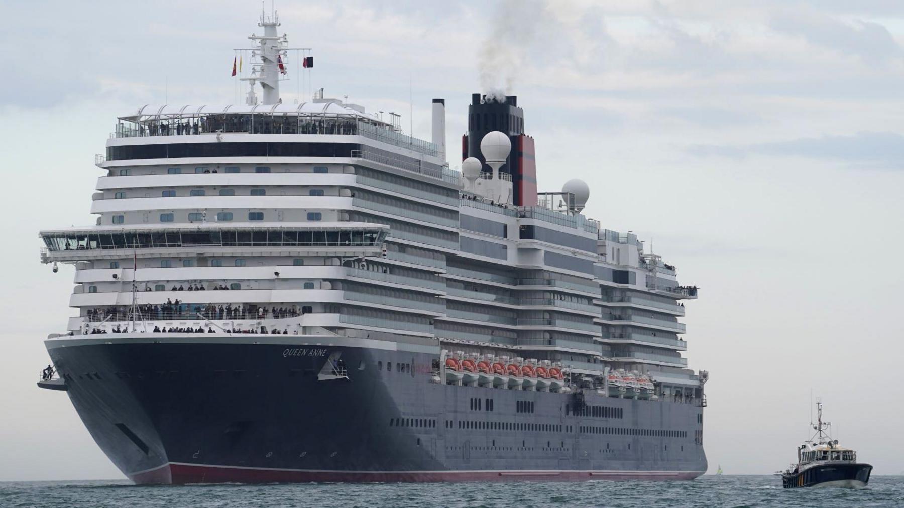 A large white cruise ship with a navy hull sails through the water. It is being accompanied into a dock by a small boat.