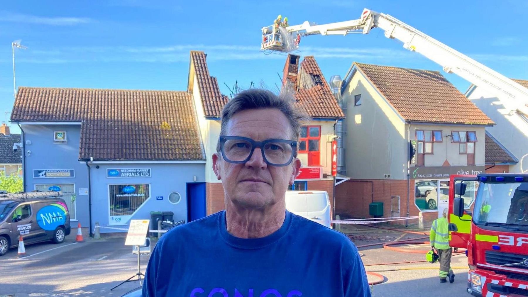 A picture of Simon Little at the scene of the fire. He smiles at the camera. He has short dark hair, blue glasses and wears a blue T-shirt. Firefighters can be seen behind him.