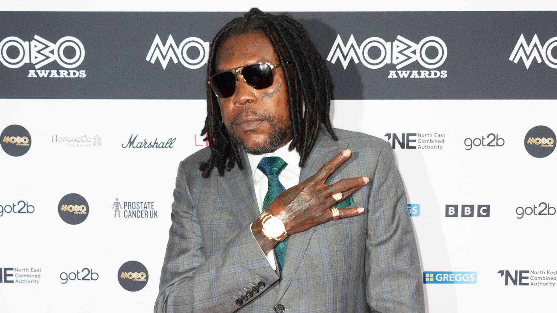 Vybz Kartel at the Mobos in Newcastle. He wears a grey suit with a white shirt and green tie, as well as dark sunglasses and a watch. He holds his right arm across his chest, his hand making an 'E' gesture. 