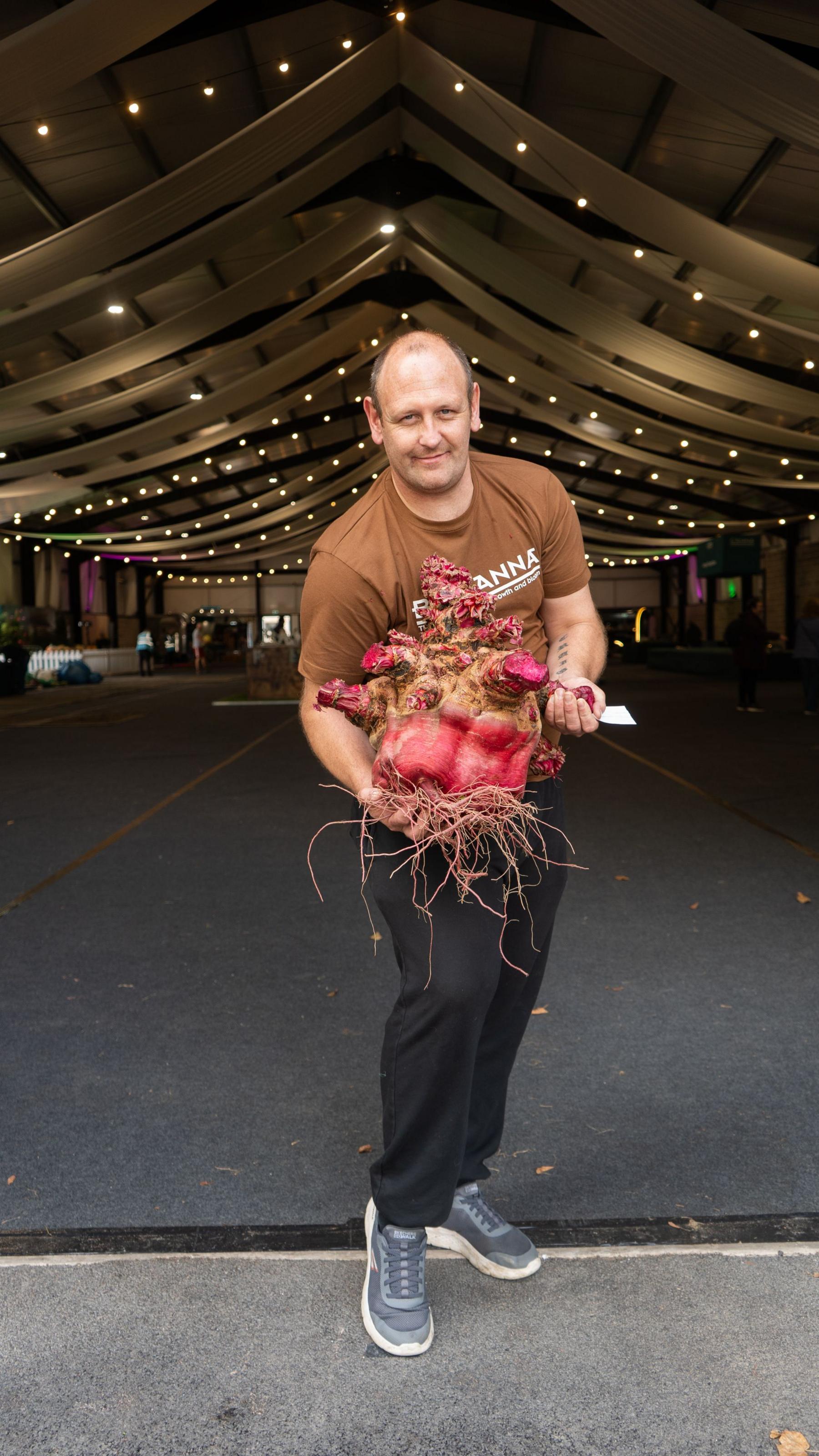 A man holds up a huge beetroot, lent on his popped knee. Large warehouse behind him, lit with fairy lights. 