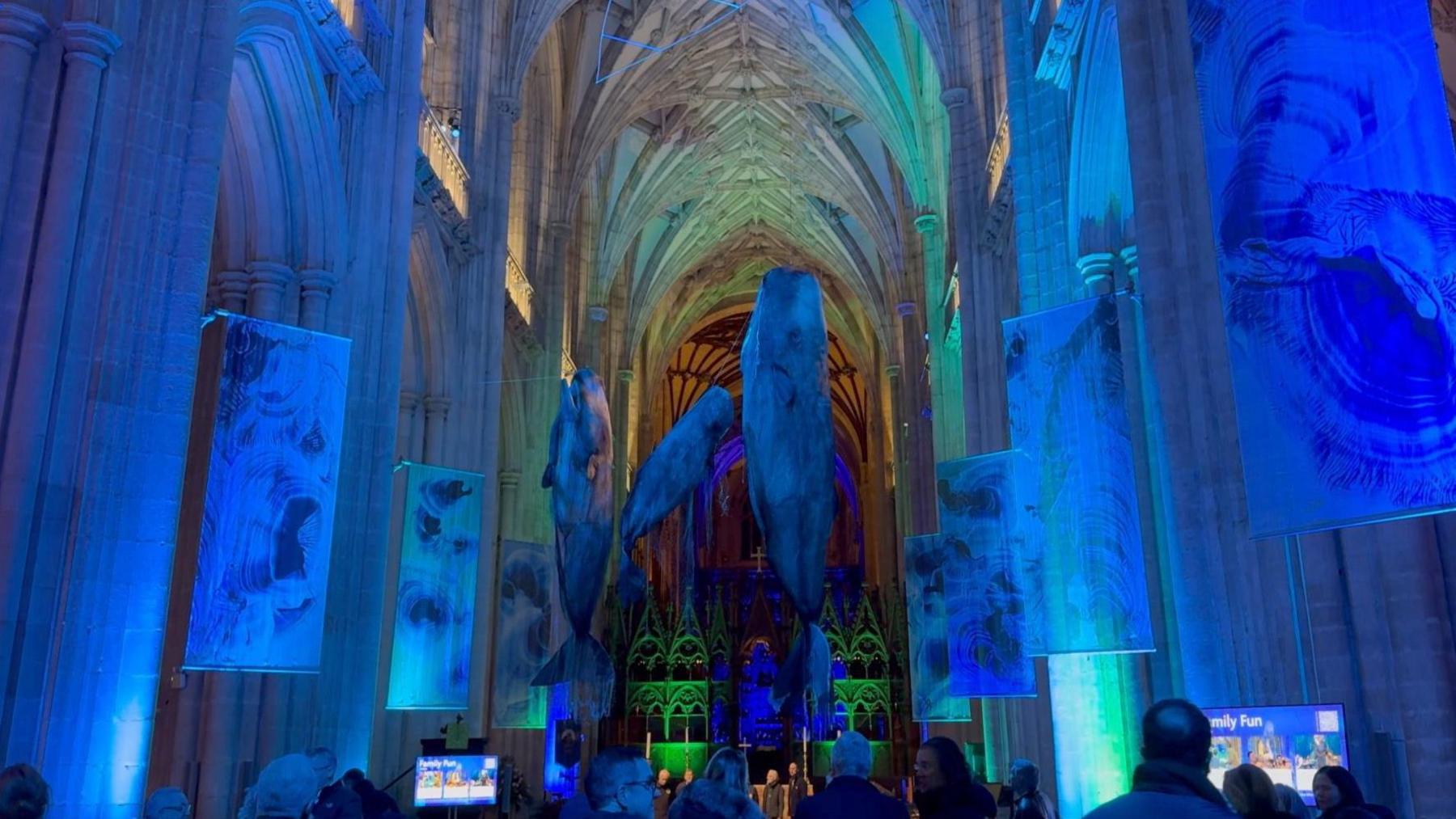 A different angle of the whales inside Winchester Cathedral's nave. Again the cathedral is dimly lit with a blue tint to it to make it feel like an immersive underwater scene. The three whales are a grey colour and there are multicoloured lights towards the back of the cathedral in green, orange and yellow.