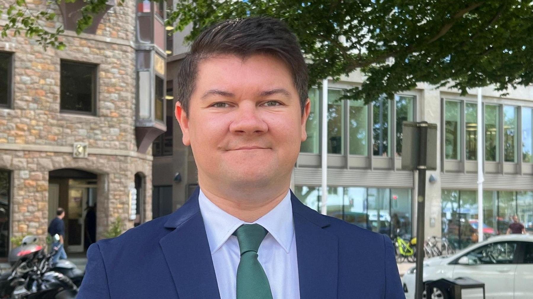 Deputy Sam Mézec is wearing a blue suit and a green tie