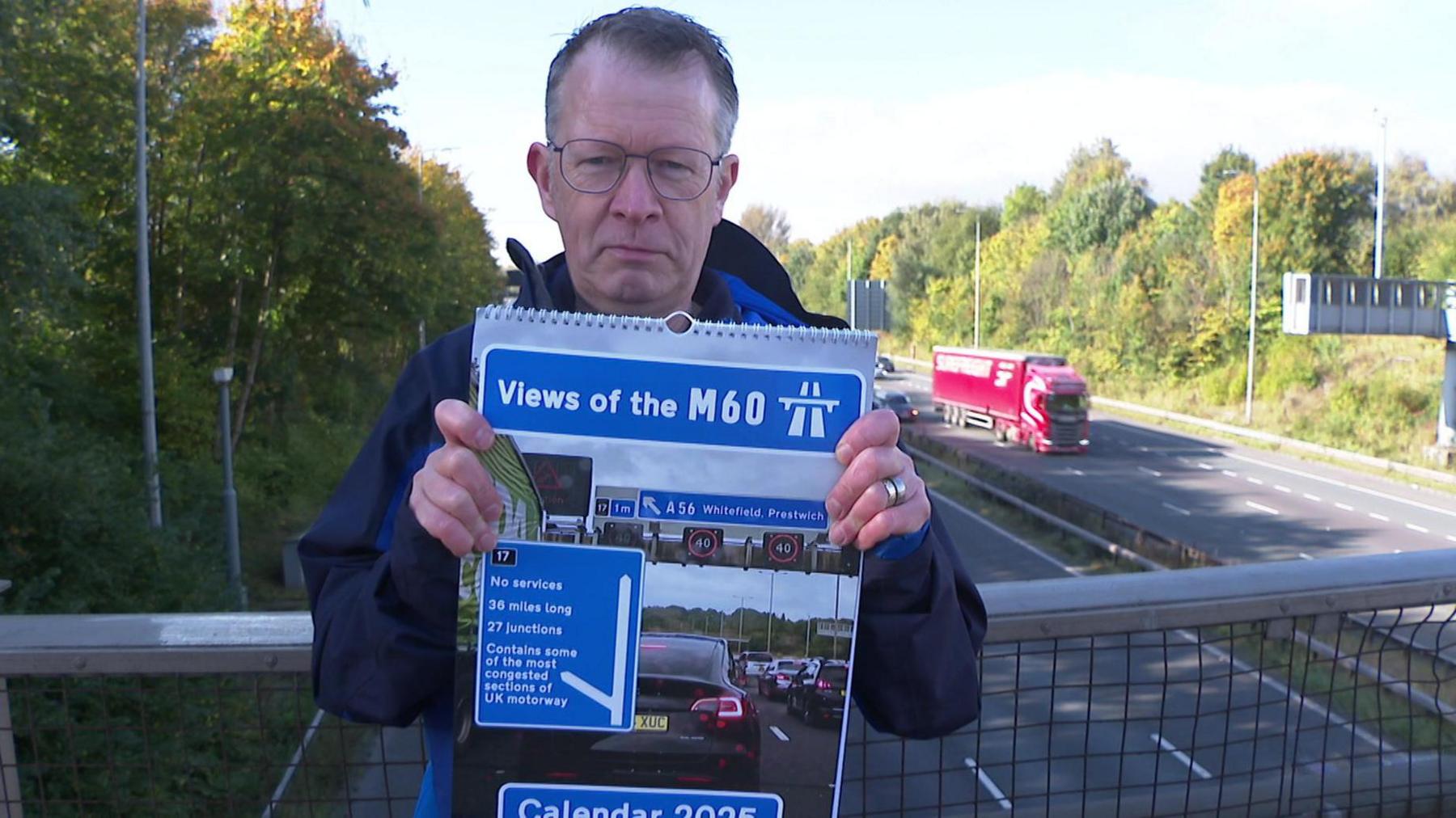 Man stood on motorway bridge with traffic passing by whilst holding copy of Views of the M60 calendar