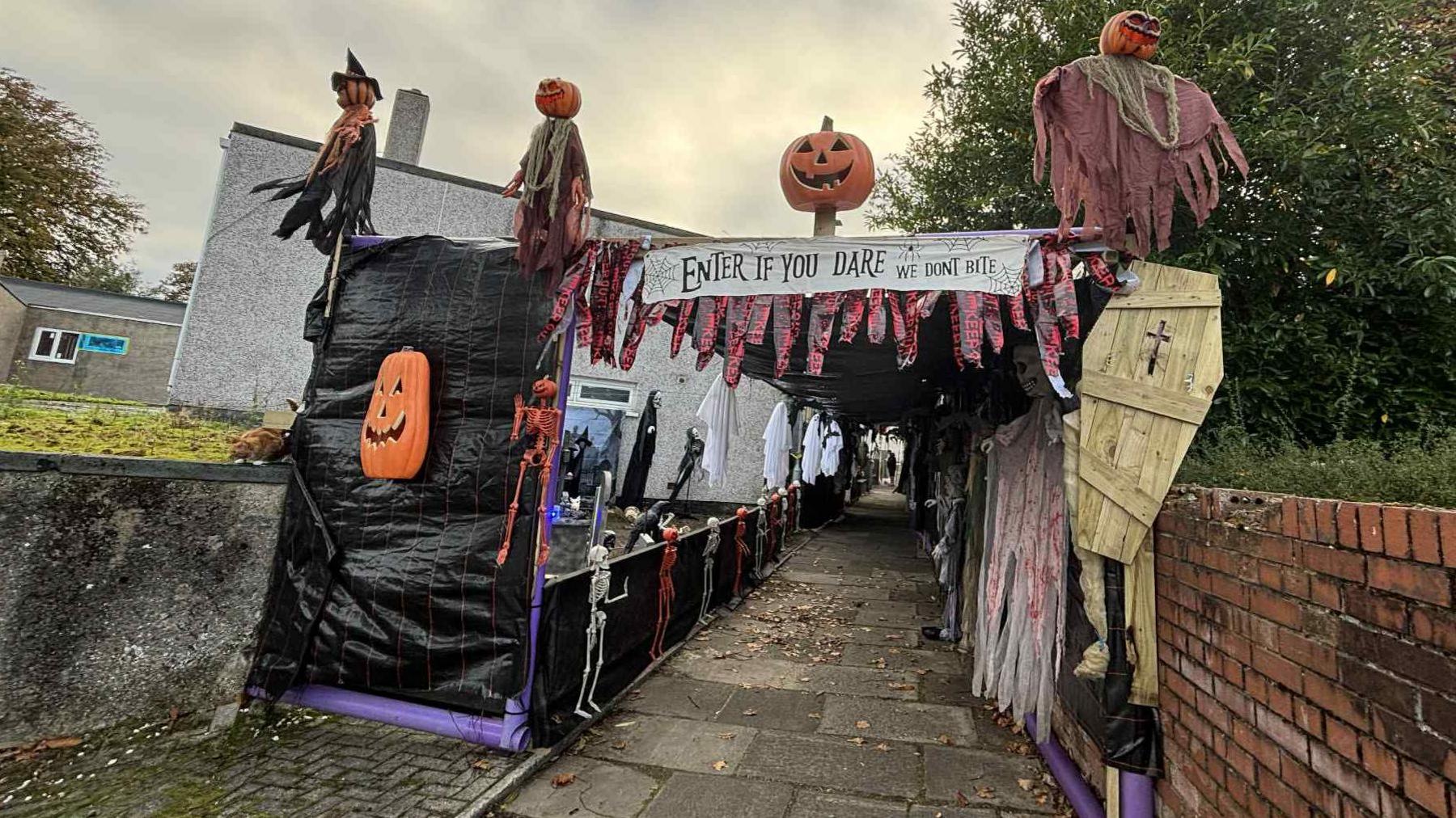 A footpath with Halloween decorations going down either side of it, on te railings and wall. A sign saying Enter if you Dare is hanging up at the entrance.