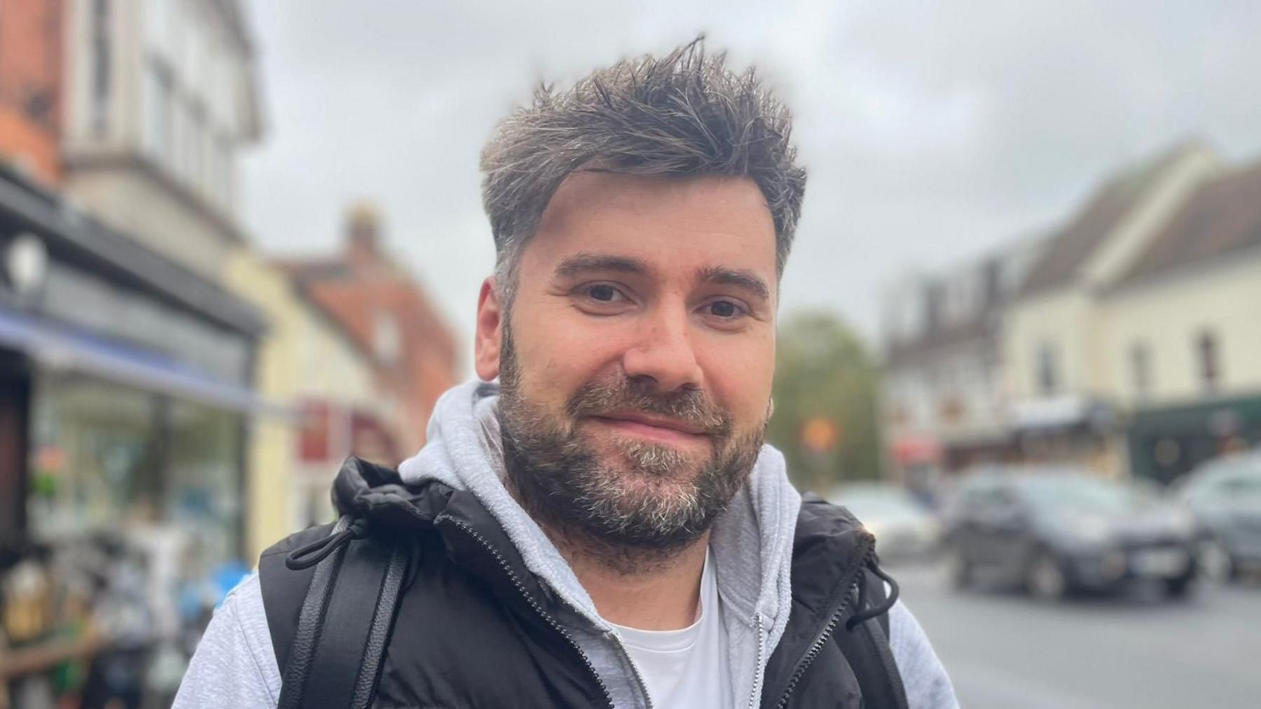 A man is smiling at camera. He has short grey hair with a grey beard. He is wearing a black padded vest over a grey hoodie and white T-shirt. He is standing outside with shops and cars behind him.