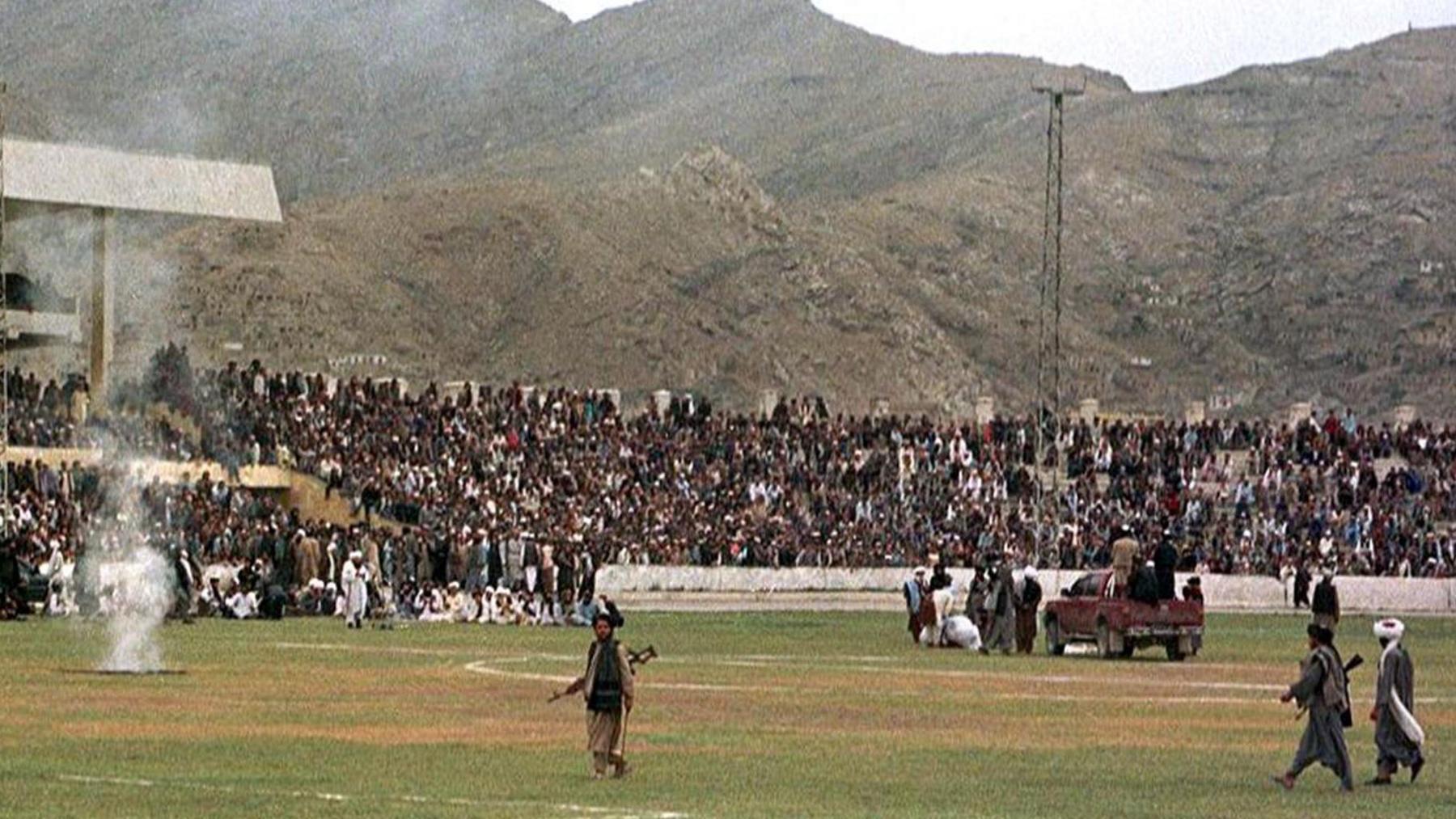 A group of Men in traditional Afghan dress hold guns as they walk across the grass in the middle of a stadium. On the right a man is hunched over on the ground next to a group of men and a pick-up truck. Crowds of people watch from the stands.