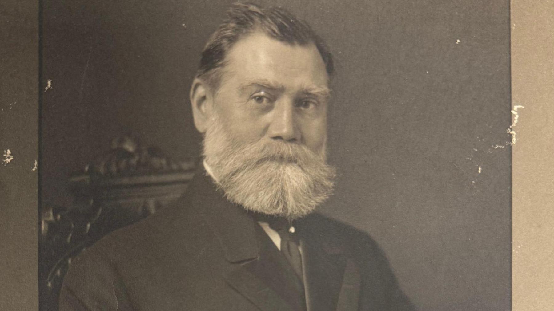 A black and white photo of Frederick Sutton, sitting on a carved wooden chair. He has a large white beard, short hair and wears a smart suit of the period.