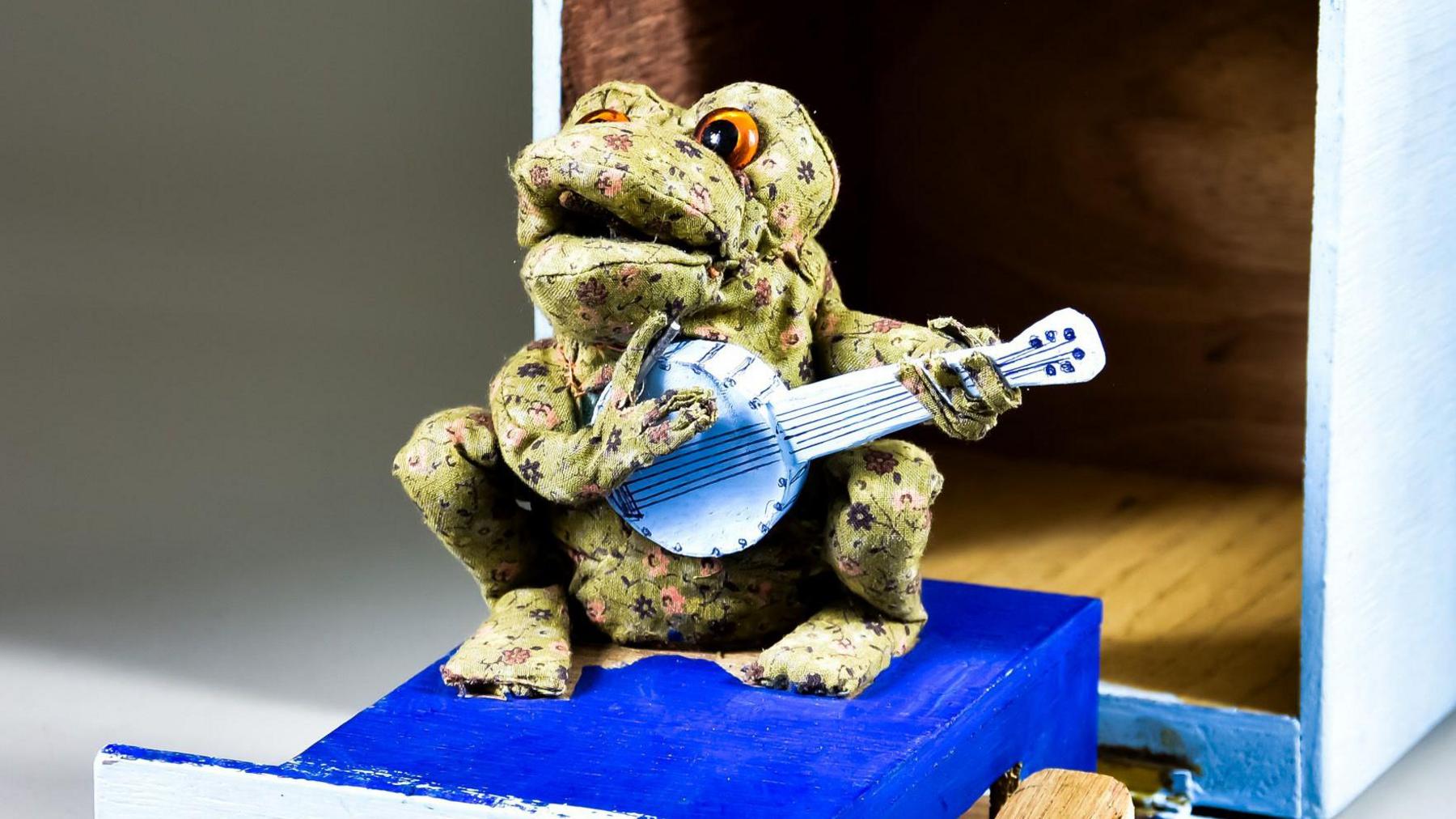 A model of Gabriel the Toad sitting on a blue platform and holding a banjo. The character has patterned green fabric covering the body, large painted eyes and is positioned beside an open blue-and-white box.