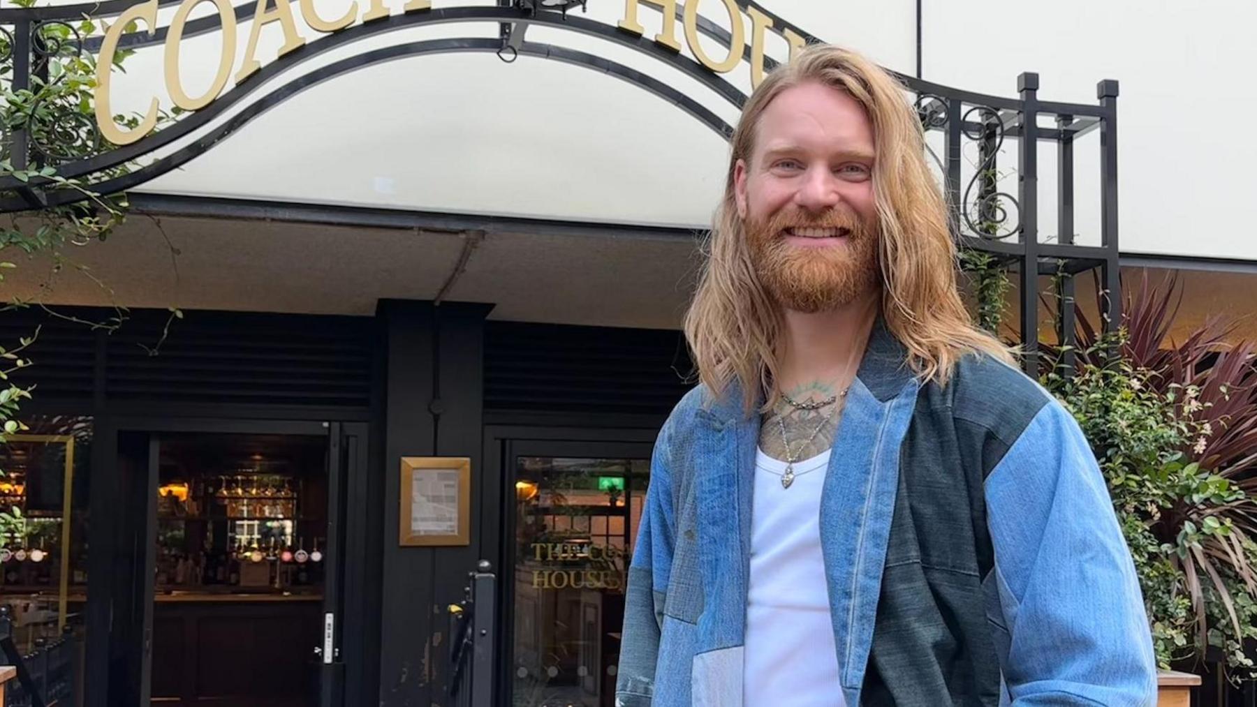 Sam is stood outside a pub, smiling down towards the camera, which is angled slightly lower than him. He has shoulder-length, strawberry blonde hair and beard. He is wearing a denim jacket with a white top underneath and two short necklaces.