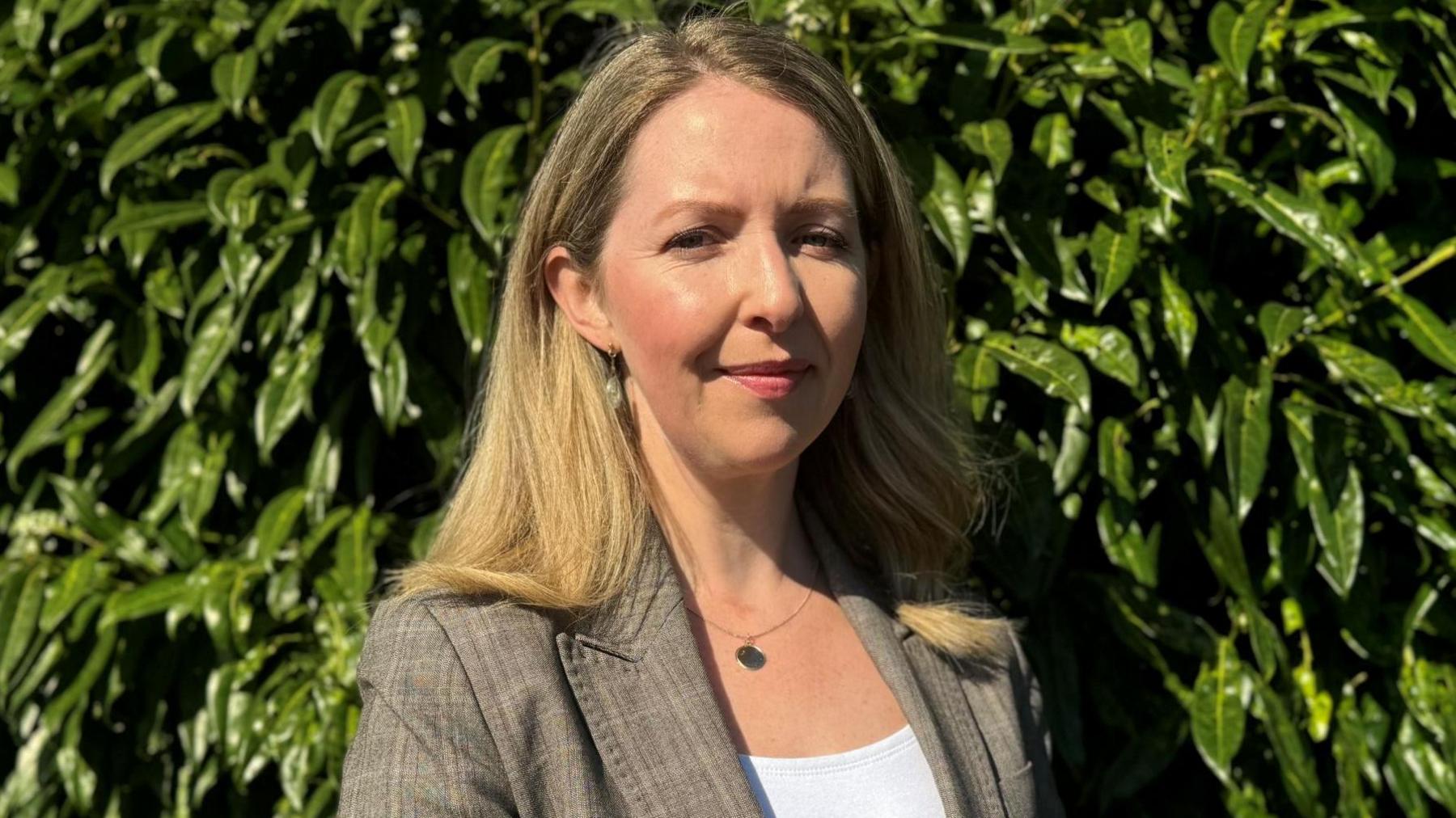 Maria McCloskey - a woman with long blonde hair stands in front of green shubbery. She is wearing a light brown blazer jacket and a white top.