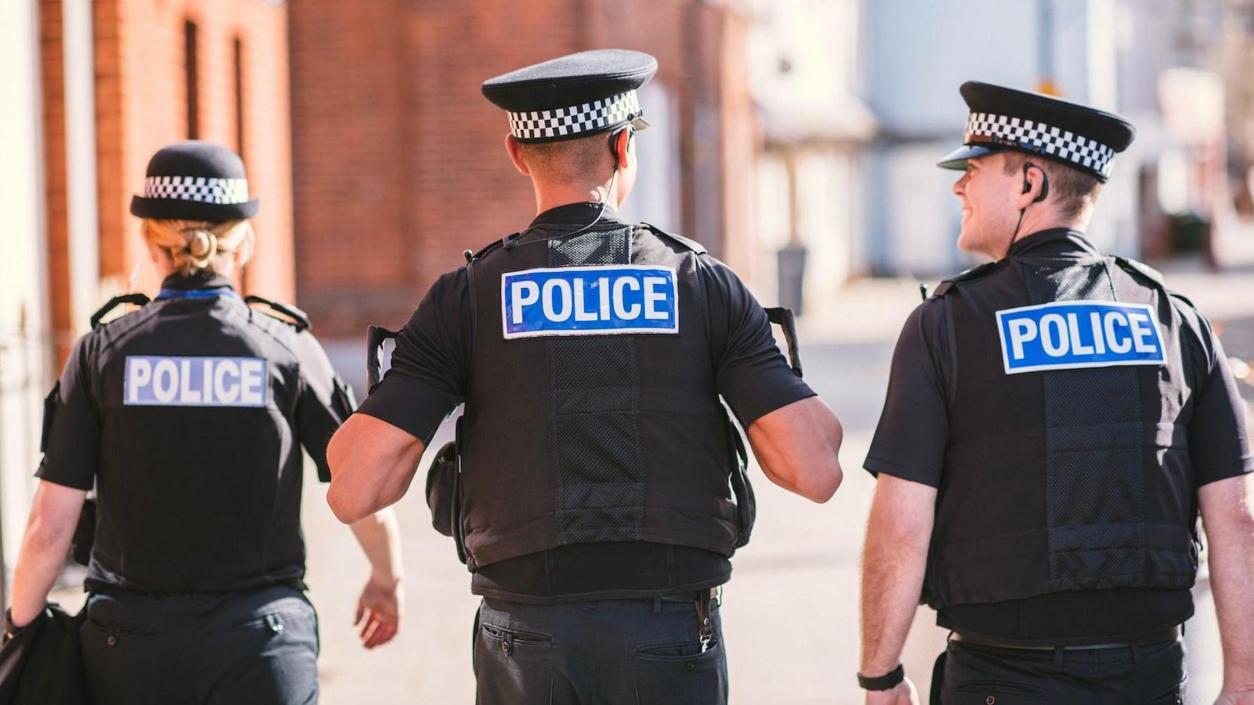 Three police officers in black uniforms and blue-and-white checkered hats walk down an urban street, facing away from the camera. Each officer wears a vest labeled 'POLICE' in bold white letters on a blue background.