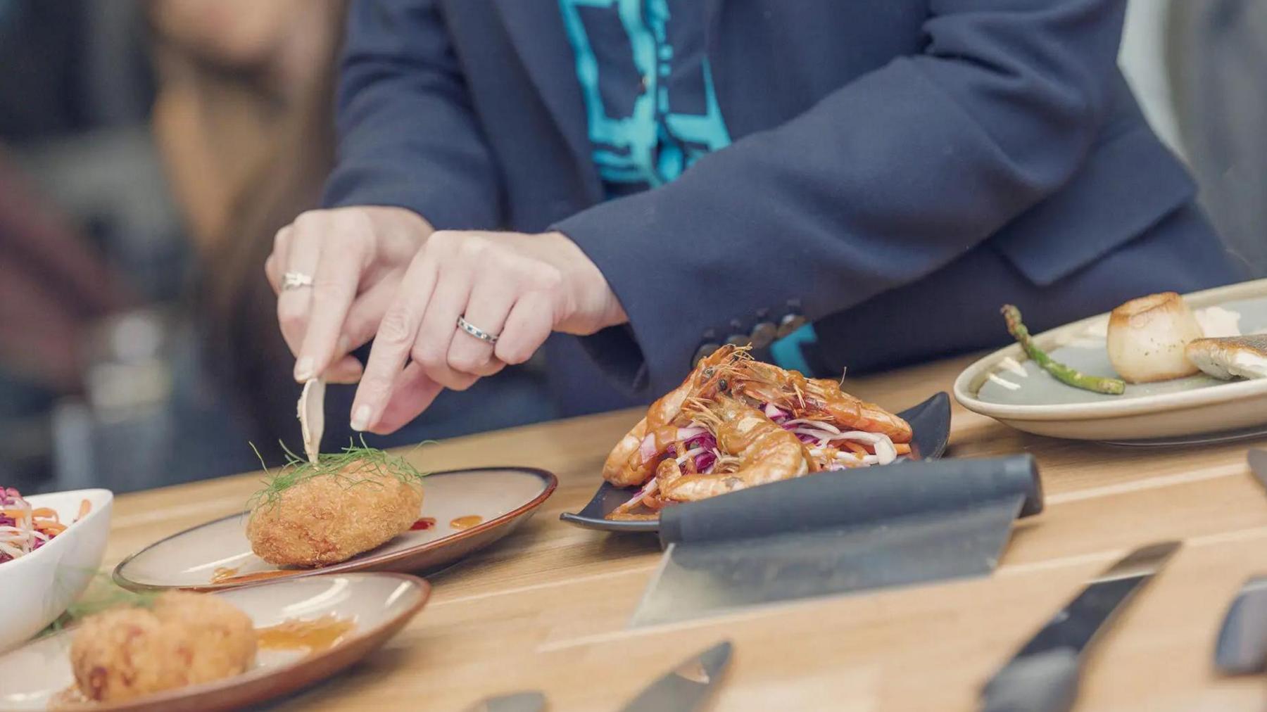 A woman cuts into samples of seafood dishes at the new school, including fish cakes, sauteed king prawns and fried scallops, all presented like restaurant dishes. 