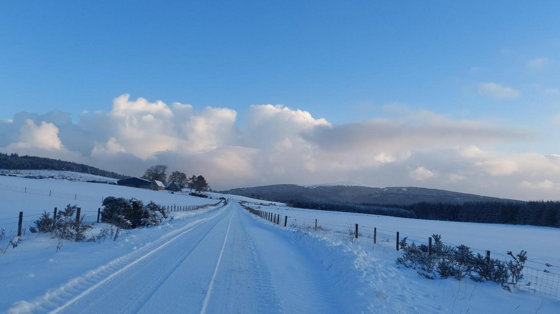 A rural road is covered in snow. Deep snow also covers surrounding fields and the roof of a farm in the distance. It is daytime and there are clouds in the sky.