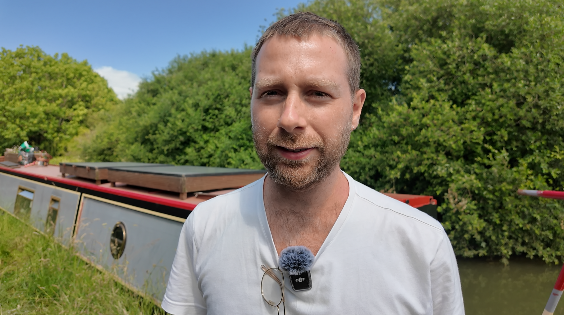 A man in a white short looks at the camera. A grey and red narrowboat floats on the canal in the background.