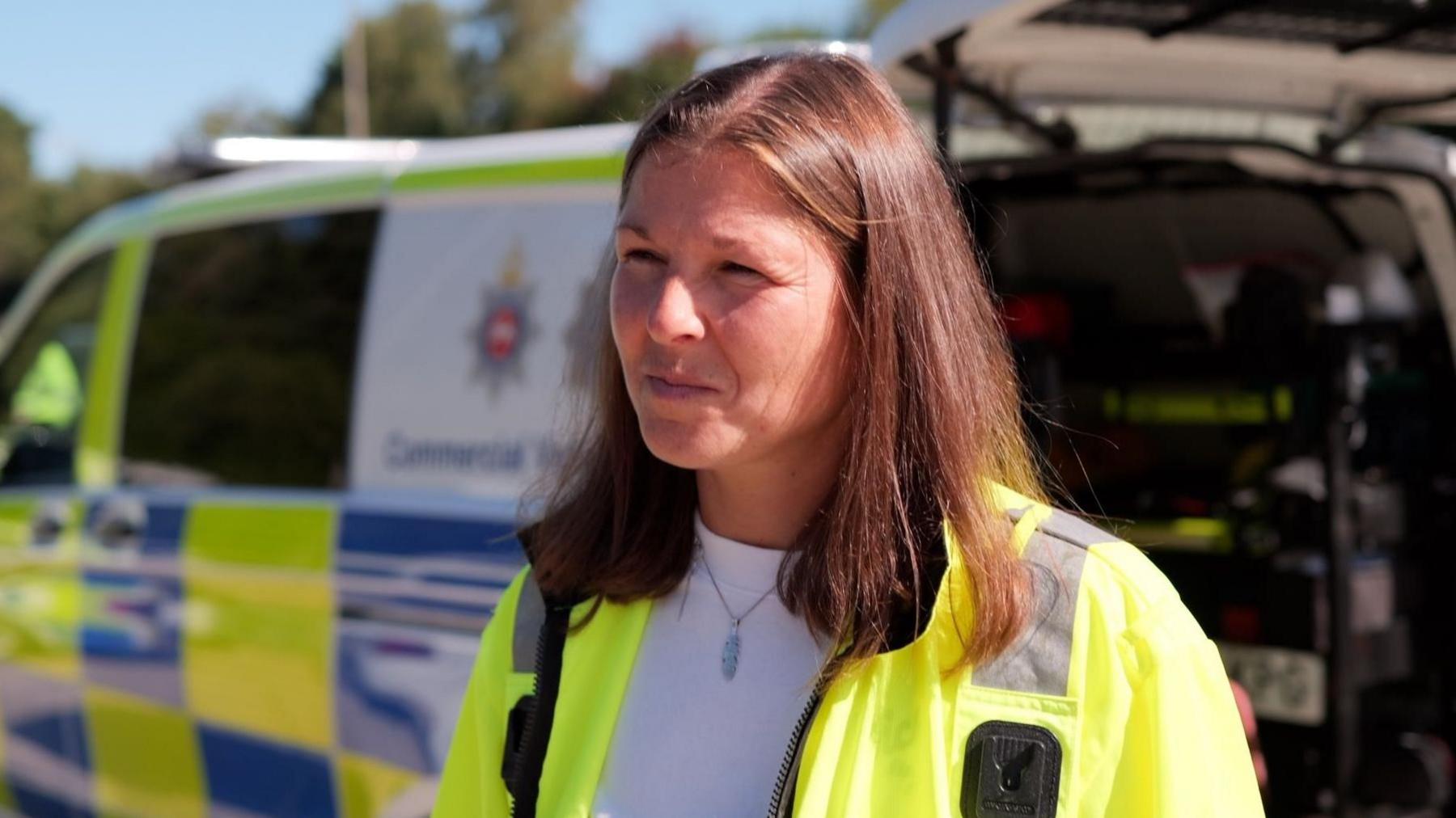 Maria Dennis, wearing a high-vis vest, stands in front of a Sussex Police van which is slightly blurred. She has brown hair just below shoulder length.