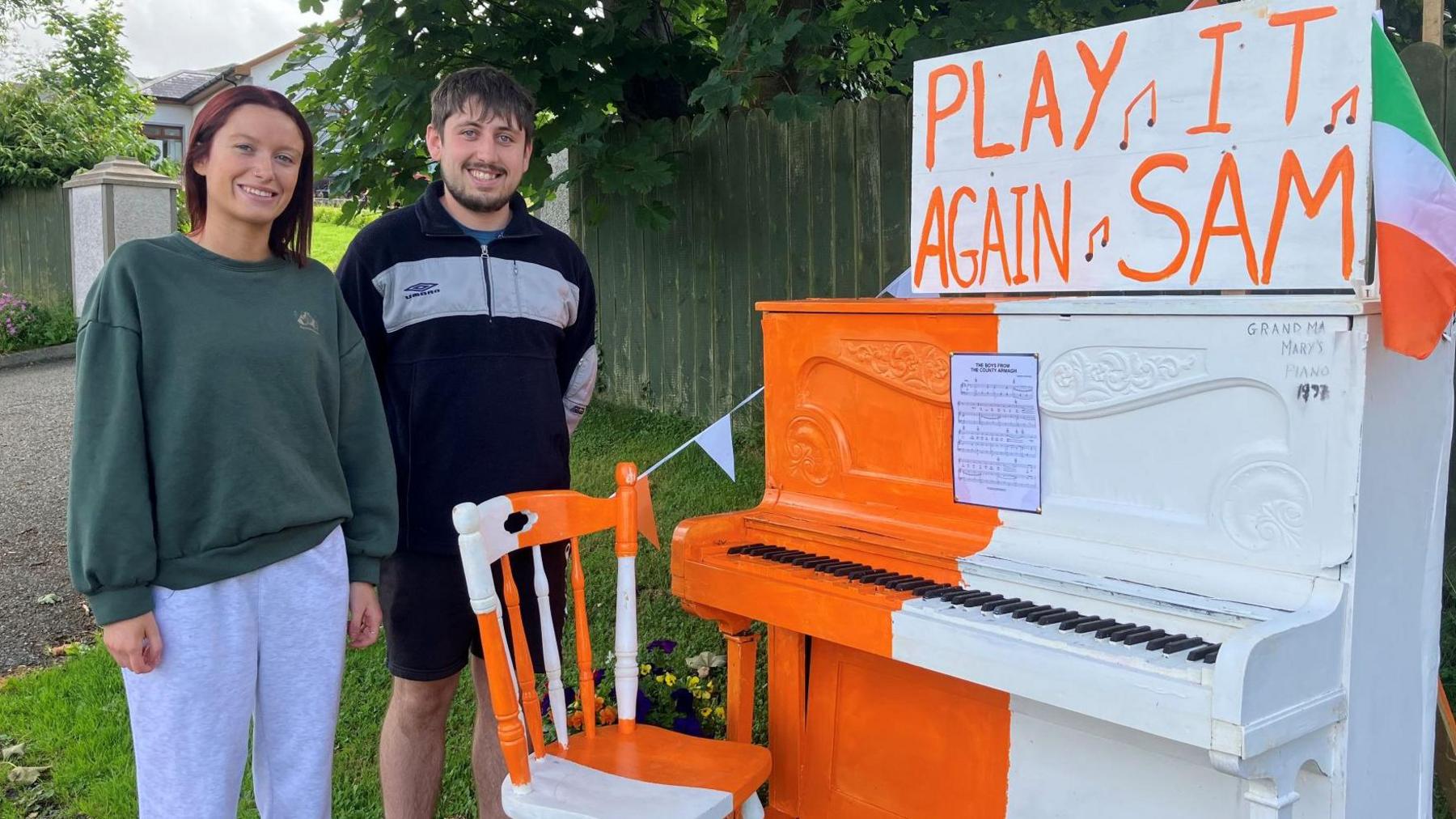 Chloe and Ogie Marks standing in front of a piano, coloured in orange and white