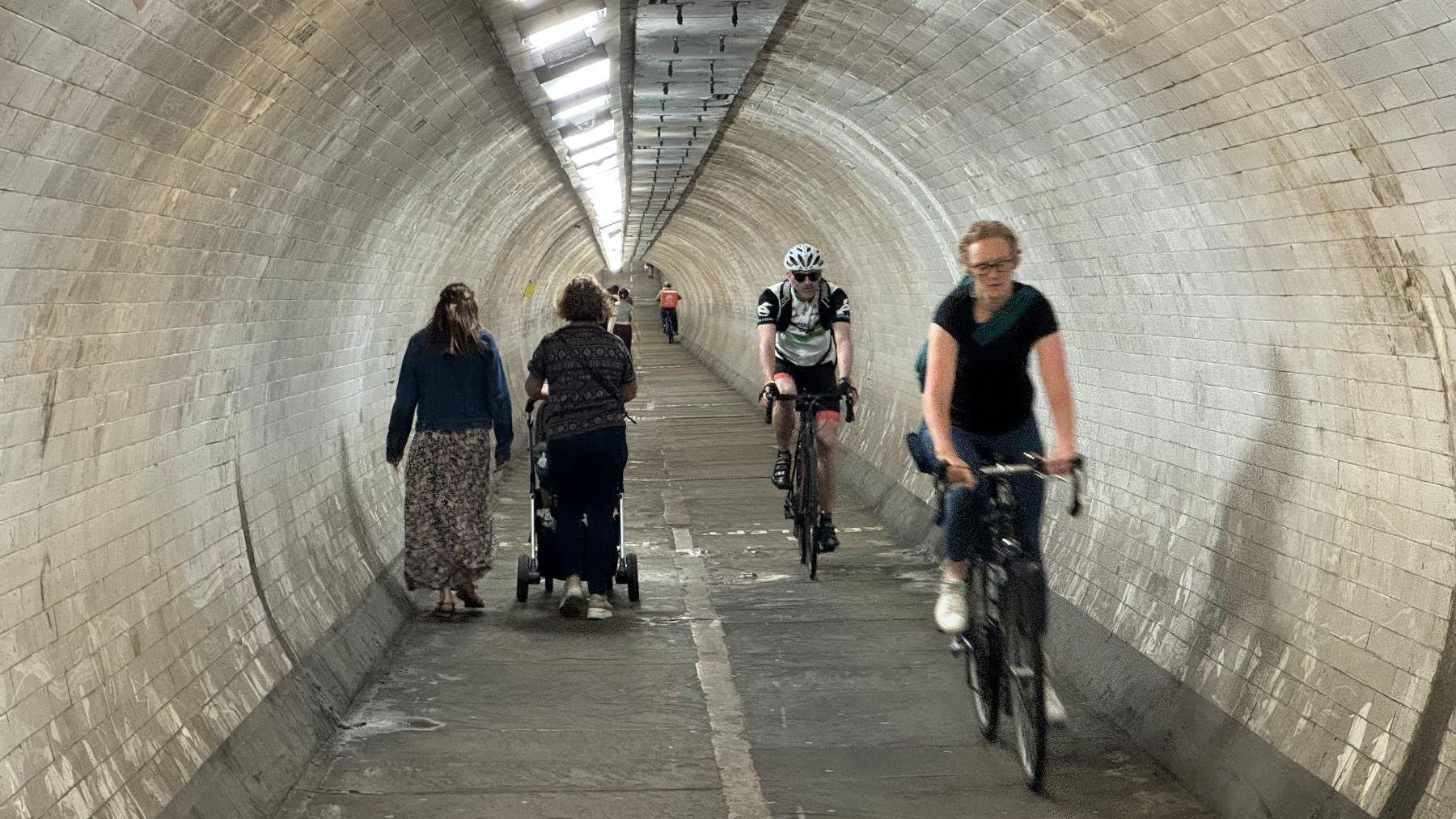 Inside one of the foot tunnels. Two cyclists are cycling towards the camera on the right, with two women, one pushing a pushchair, facing away on the left. More pedestrians using the tunnel are visible in the background.