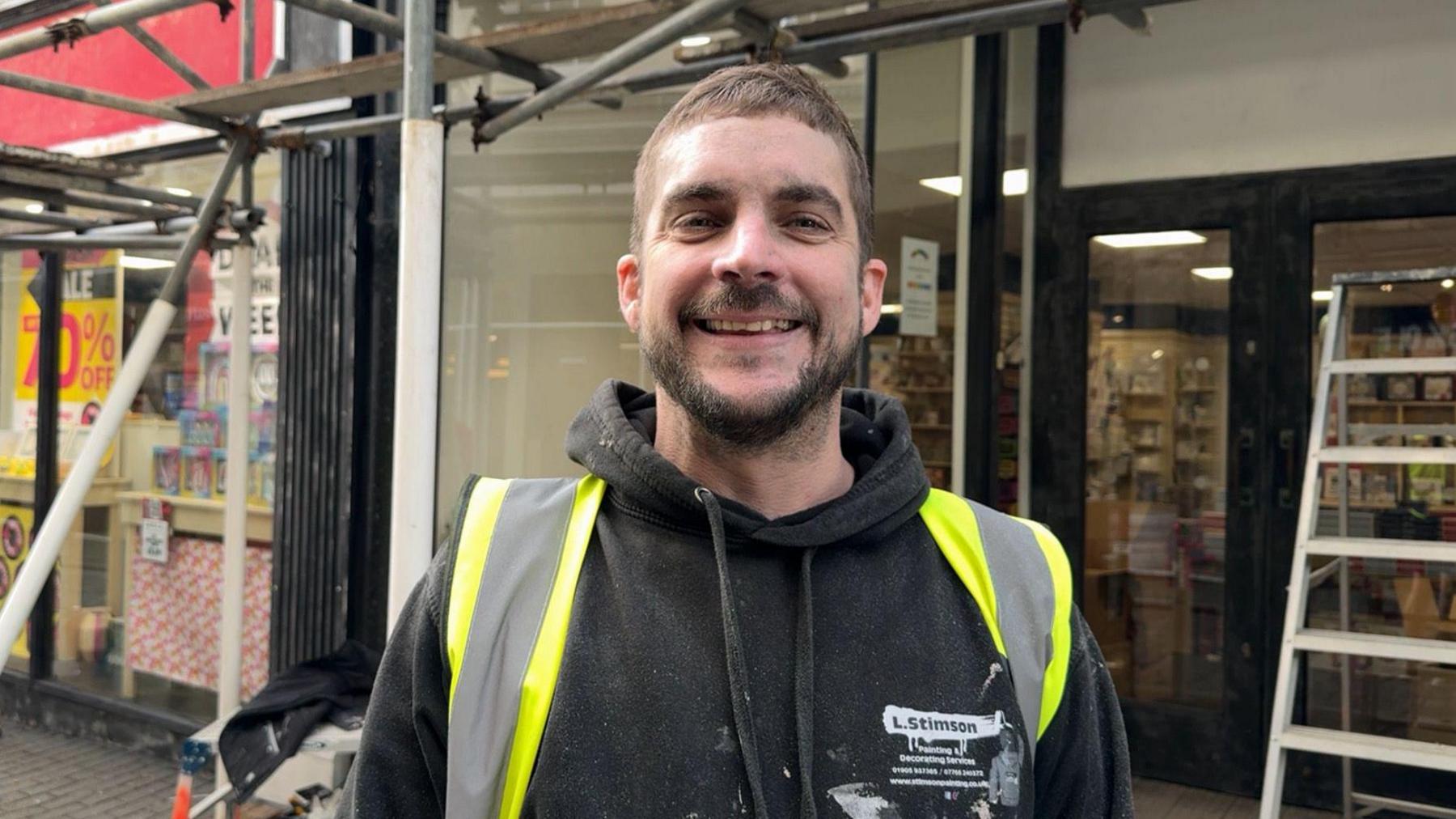 Man in his 30's with a short hair cut and a beard. He is wearing a black hoodie and a high-vis jacket.