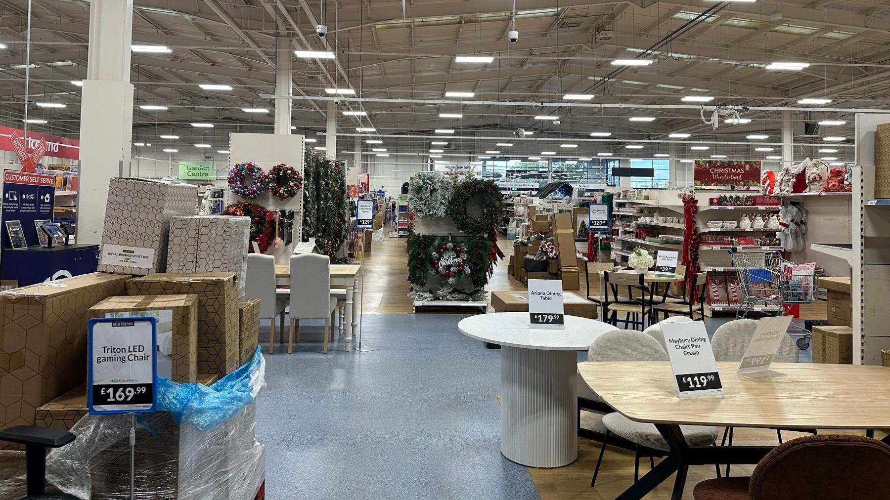 Inside the store, which has a huge warehouse style roof with multiple ceiling lights. In the foreground there are boxes are piled up and tables and chairs for sale. Further back, Christmas decorations and wreaths are on display, and there are long shelving units stretching into the distance. 