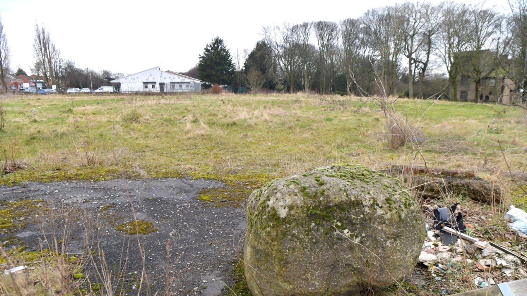 A boulder in the foreground and green scrubland with a white building (the swimming pool) in the distance to the left and part of Bolling Hall Museum to the right. 