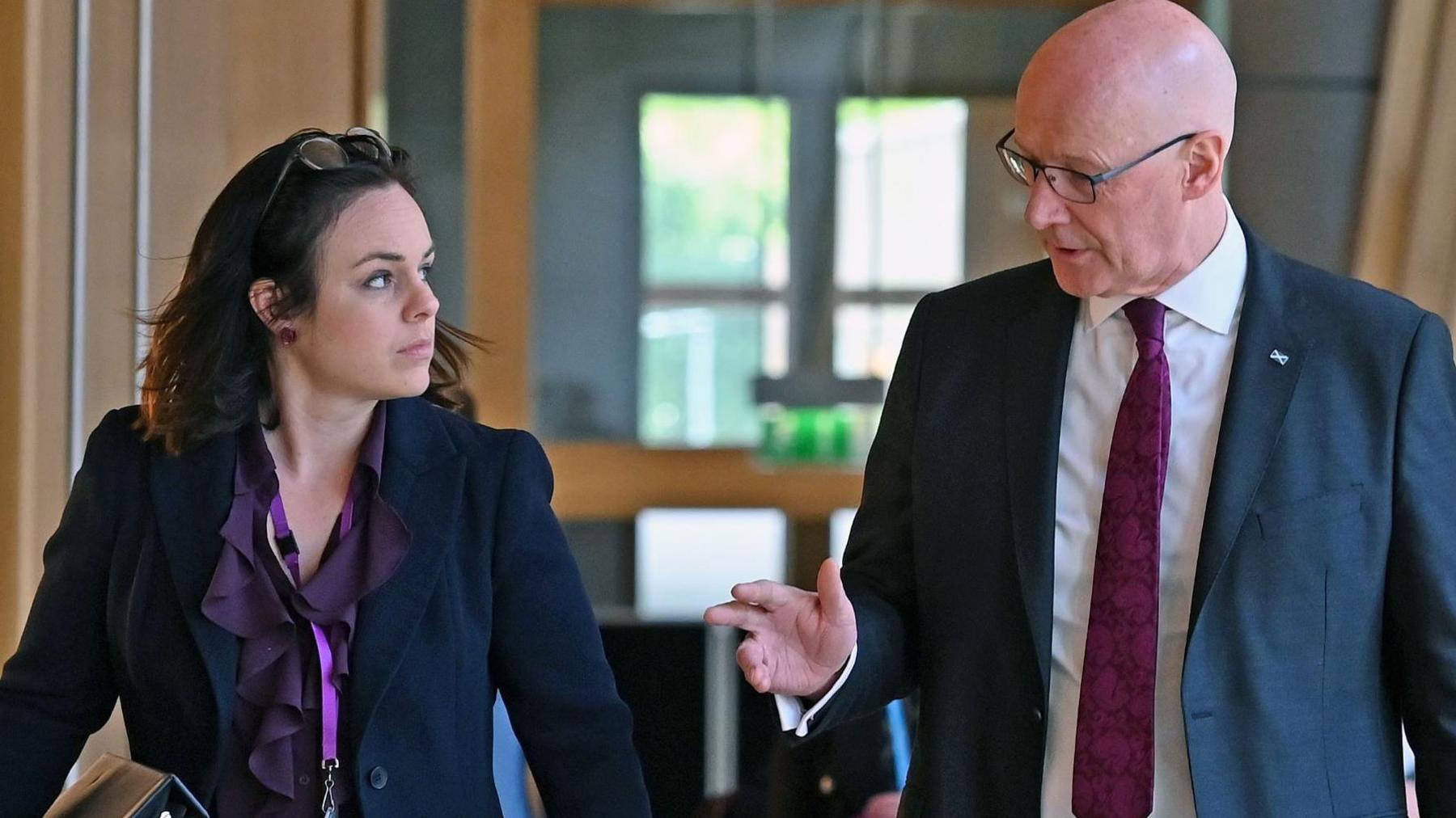 Kate Forbes and John Swinney, who is bald and wearing glasses, chatting while walking along a corridor at Holyrood. He is wearing a navy suit, white shirt and purple tie. She is wearing a navy trouser suit and a purple blouse. She is balancing her glasses on her head and has purple lanyard round her neck.