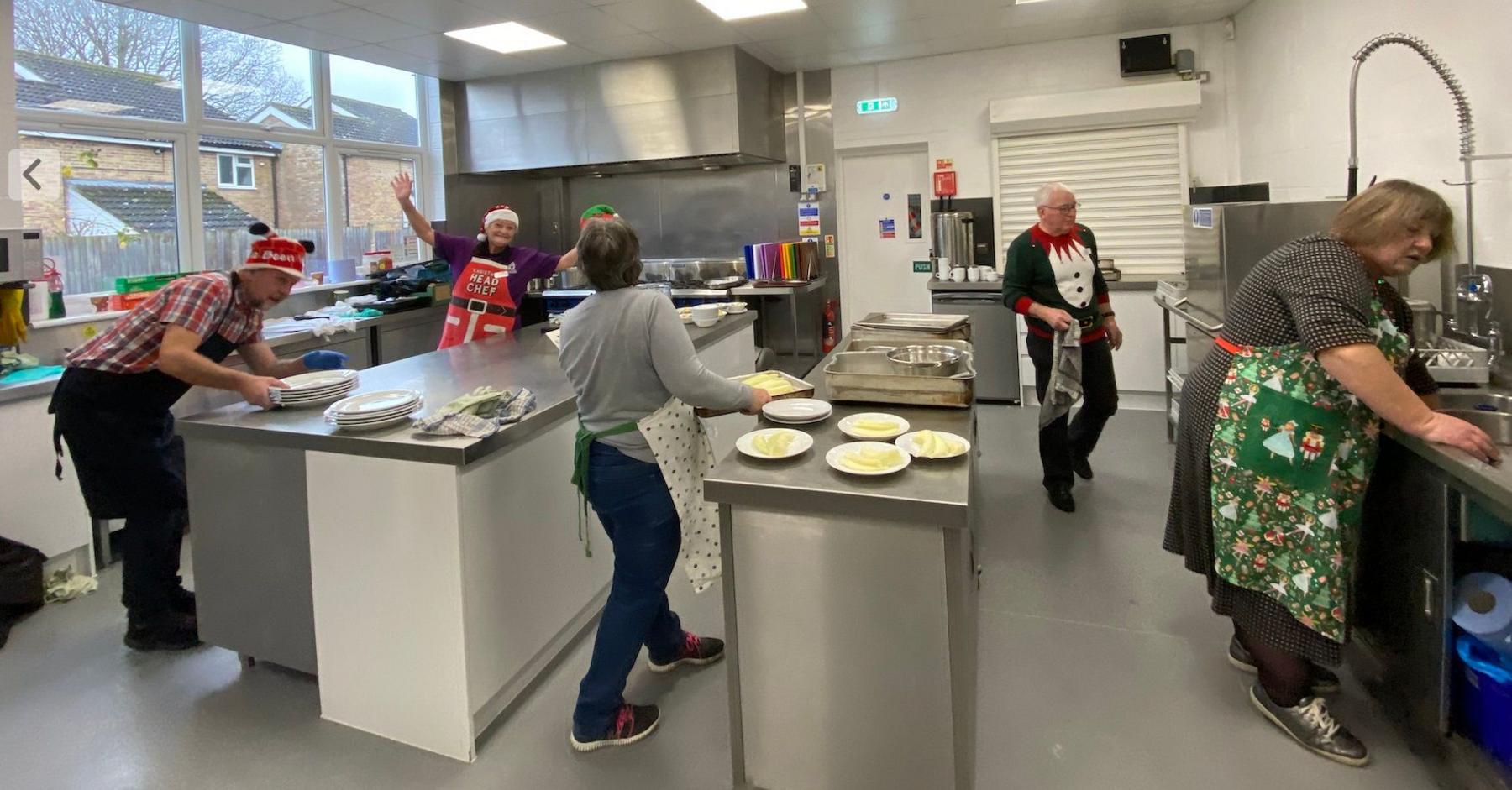 A group of volunteers working to prepare the Christmas Day lunch on Christmas Day, 2024. They can all be seen at different kitchen stations preparing food. They are all wearing Christmas themed aprons and jumpers.