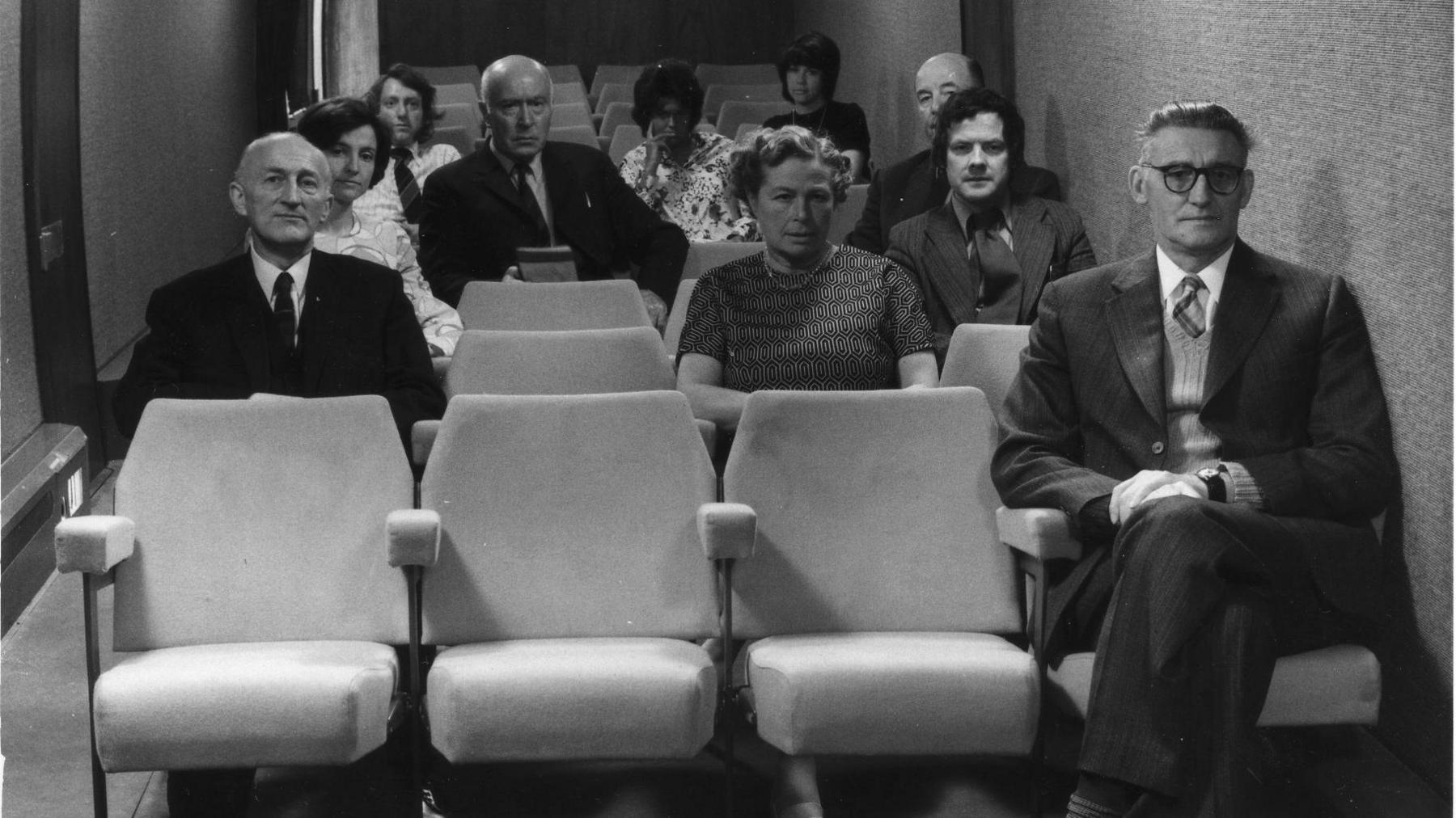 An old black & white photo from 1975, looking down a train carriage, which has cinema seats rather then usual seating for travelling. Around of the third of the seats have people in them, all looking at the camera. Some men in suits, women in dresses.