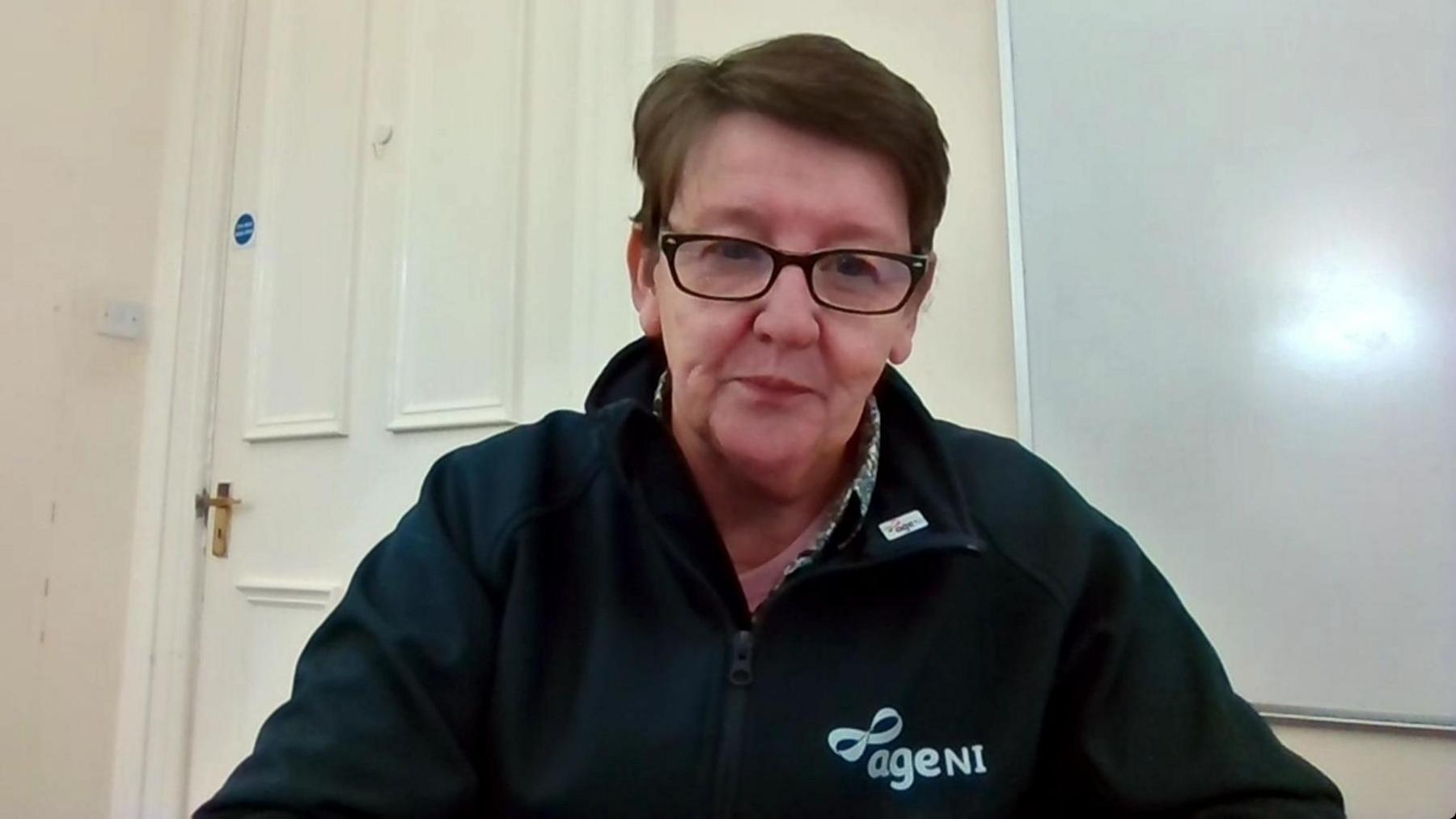 Age NI chief executive Linda Robinson on a Zoom call.  She has short, brown hair, glasses and is wearing a dark Age NI branded fleece over a patterned blouse. 