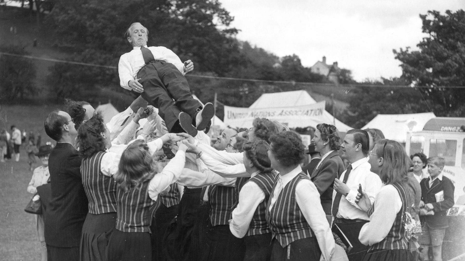 Arweinydd côr yn cael ei daflu i'r awyr a'i ddal gan weddill yr aelodau