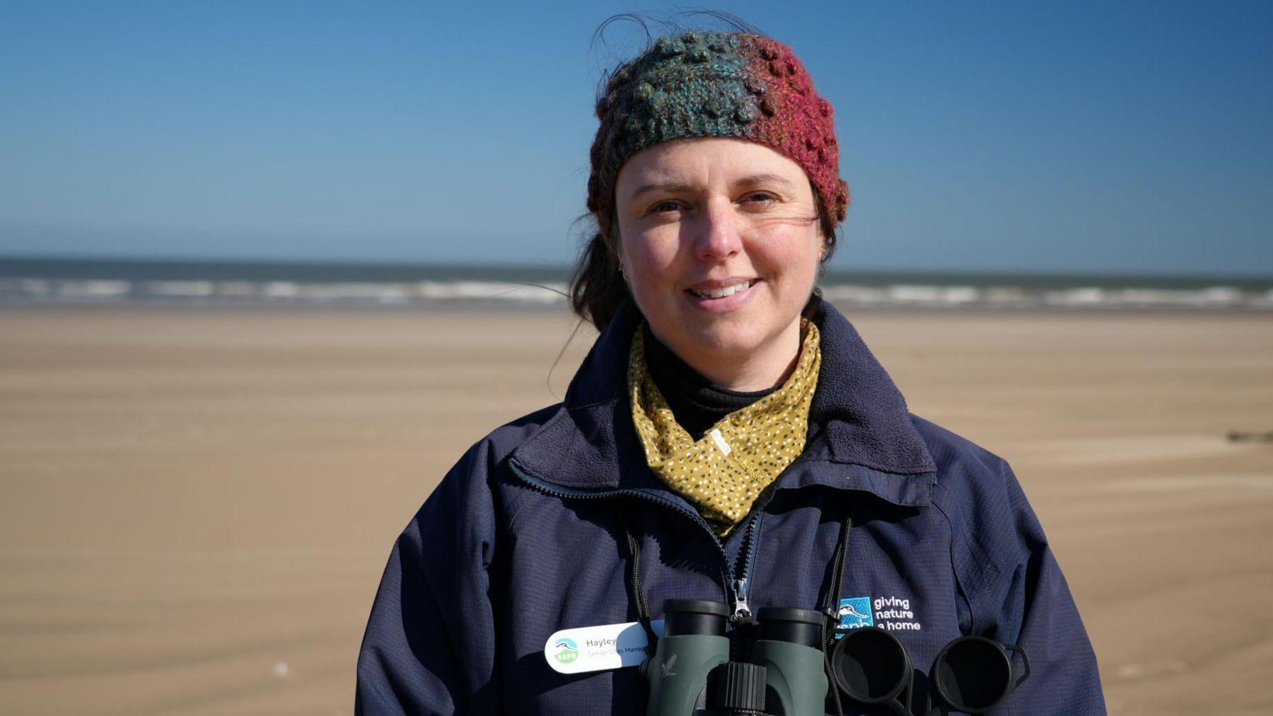 Hayley Road wearing a multi-coloured hat, yellow spotty neck gaiter and a navy blue jacket. She is stood on a sandy beach below a blue sky. Her brown hair is tied back and she is smiling