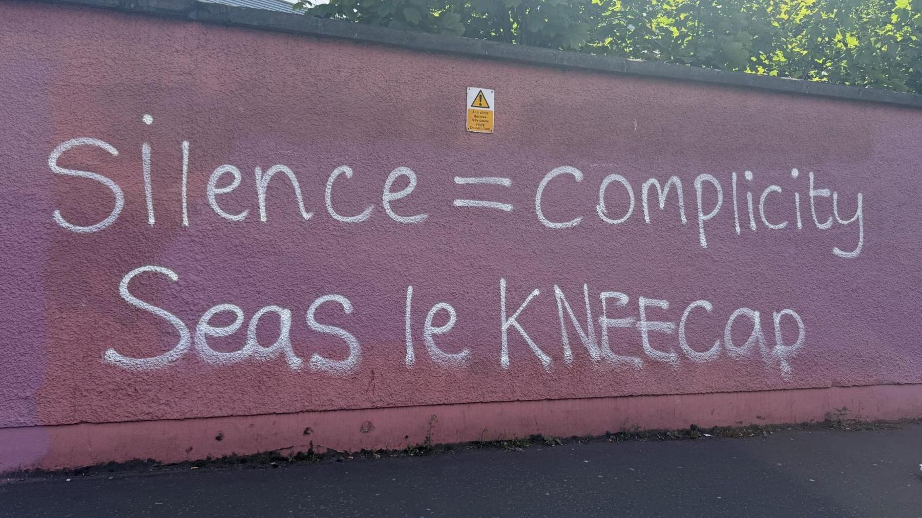 In white spray paint, 'Silence = complicity/Seas le Kneecap [stand with Kneecap in Irish]' is written on a dark pink wall. 