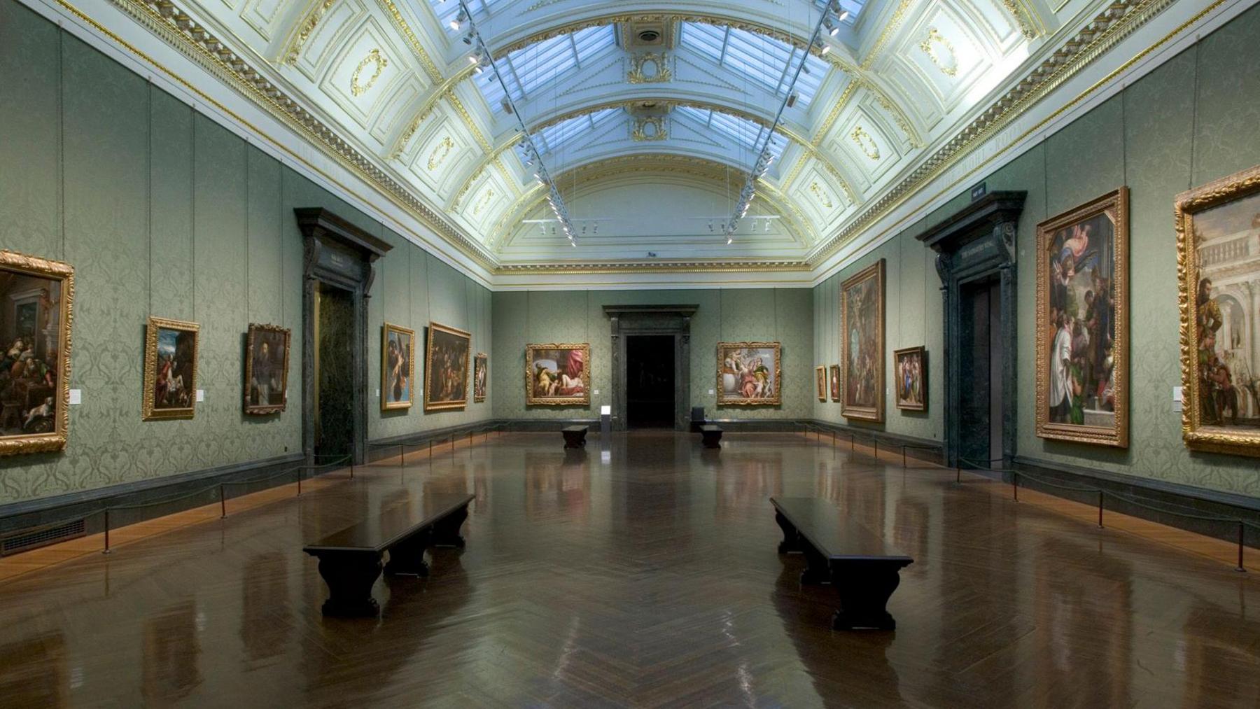 A large room in a gallery with oil paintings hung on the light green wallpapered walls along either side and benches spaced out along the hall. The hall is otherwise empty and the dark wooden shiny floorboards are in a herringbone pattern. It has a large decorative glass dome running the length of the hall with gold paintwork.