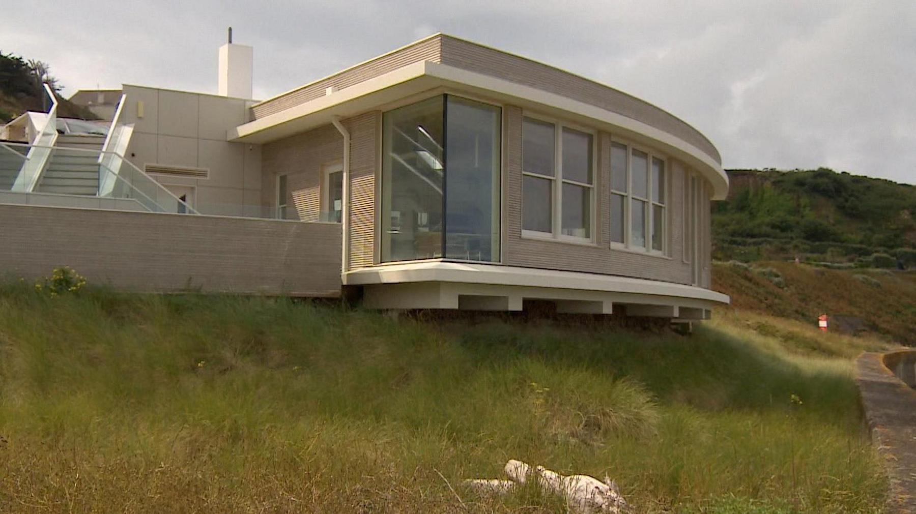 A modern one story cream building with a large window just out over grass.