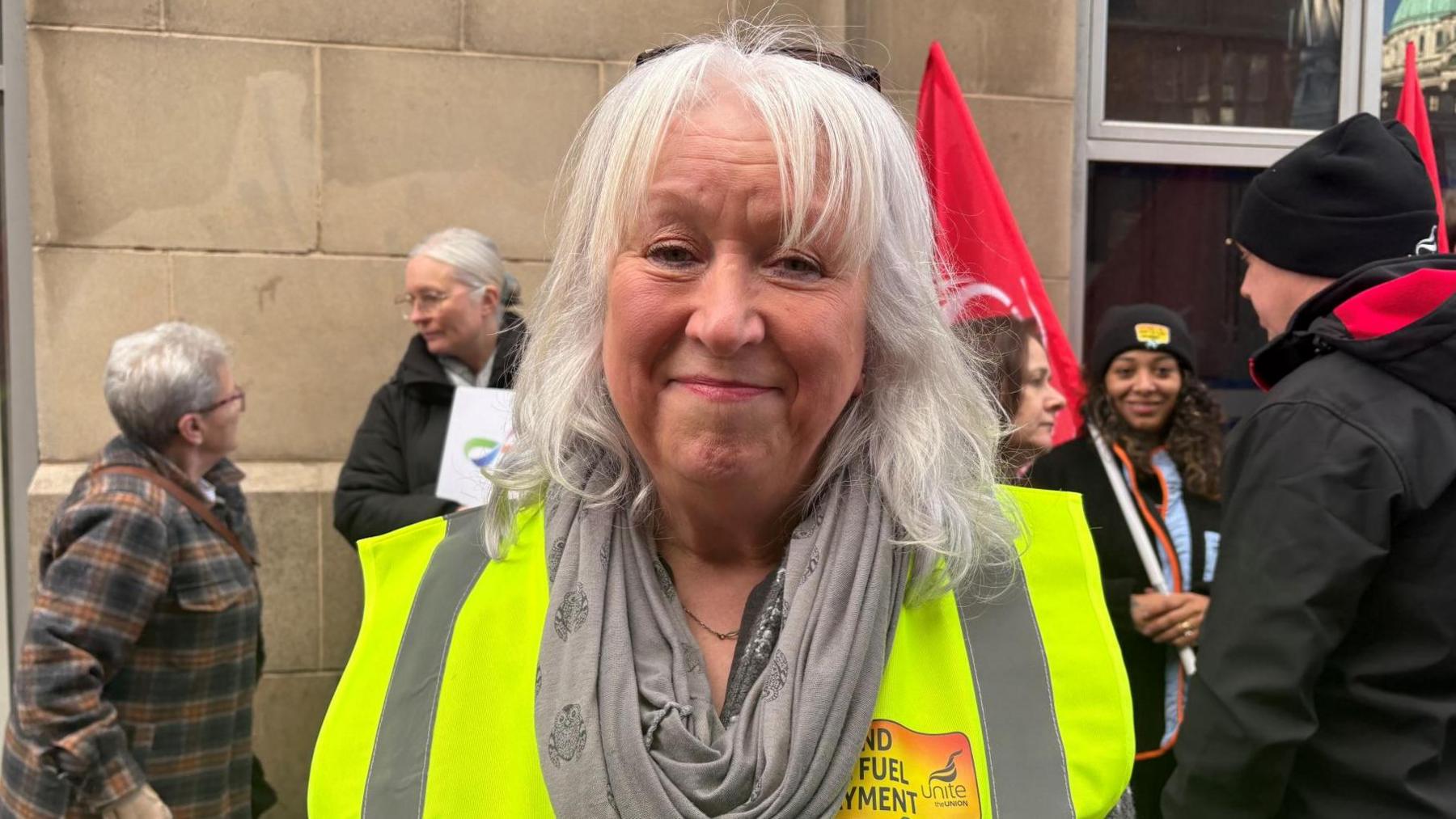 Dympna McGlade - a woman with shoulder-length grey hair is smiling at the camera as campaigners hold flags and talk in the background. She is wearing a yellow hi-vis jacket and a grey scarf.