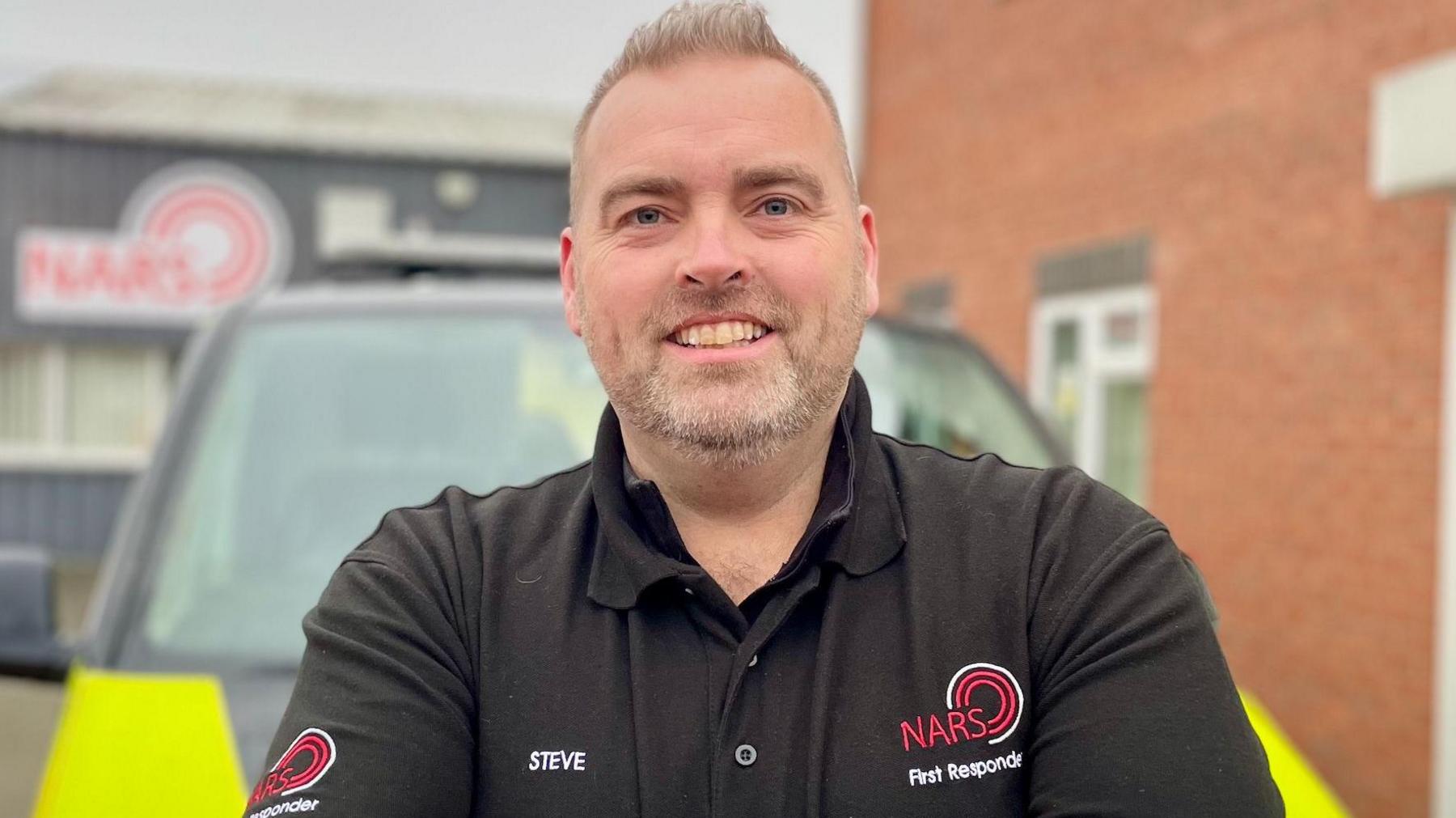 A man with short hair and stubble smiles at the camera and wears a black NARS polo shirt with his name on it. In the blurred background you can see a hi-vis vehicle