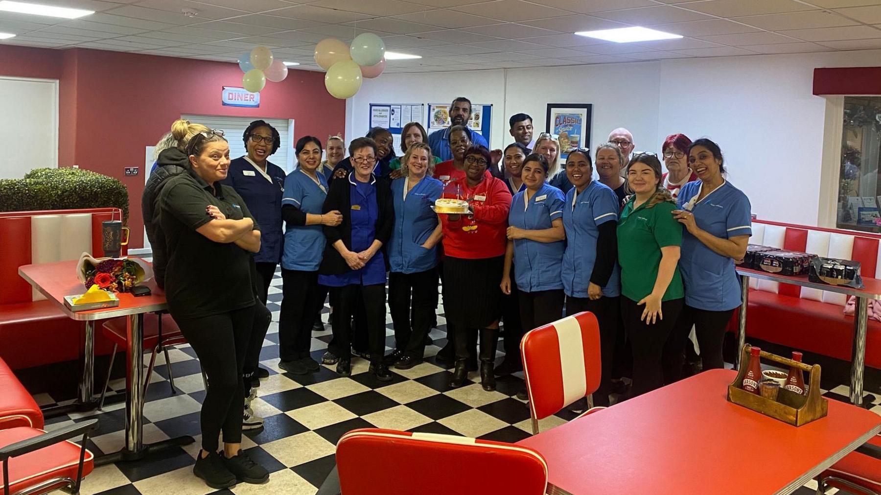 Staff have gathered together at Wrottesley Park nursing home in Wolverhampton. Some are wearing blue uniforms. There are chairs and tables in the communal area and there are balloons on the ceiling.