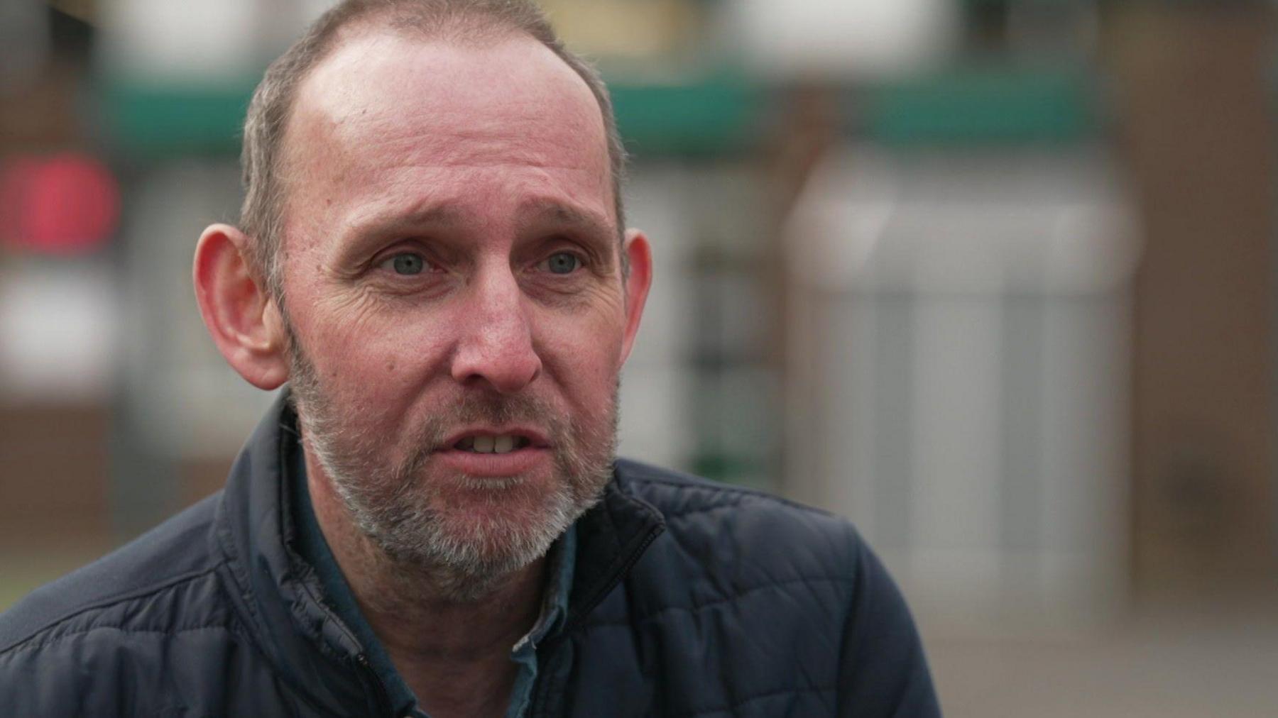 A man in his 40s with receding hair, wears a navy jacket and sits outside in a school playground, looking off from the camera 