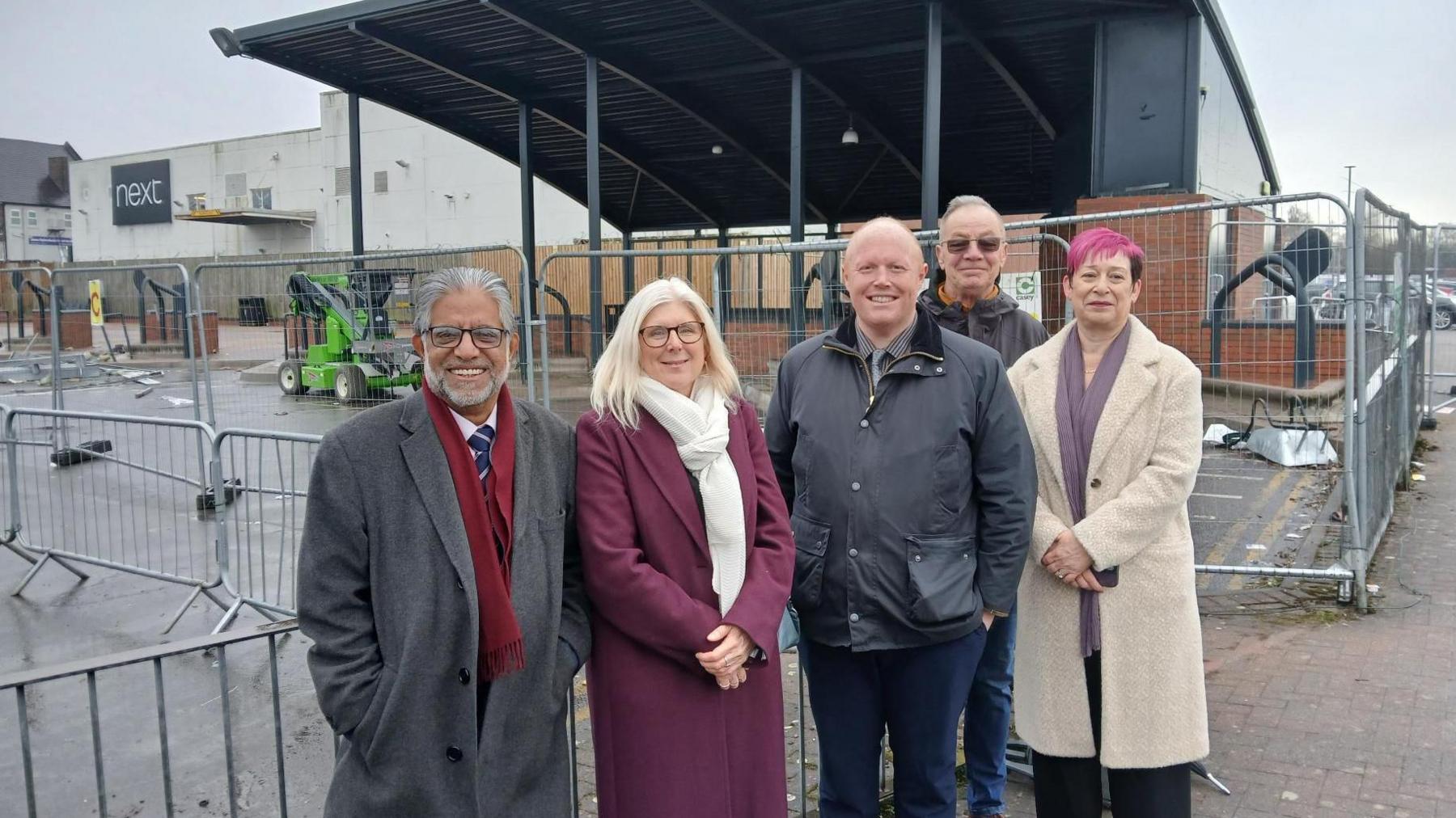 Five people stand in front of railings around a shelter at a bus station. There are three men and two women in the group, all of whom smile at the camera. The silver railings surround a tall black overhead canopy, and a white building with a black Next sign is behind the shelter.