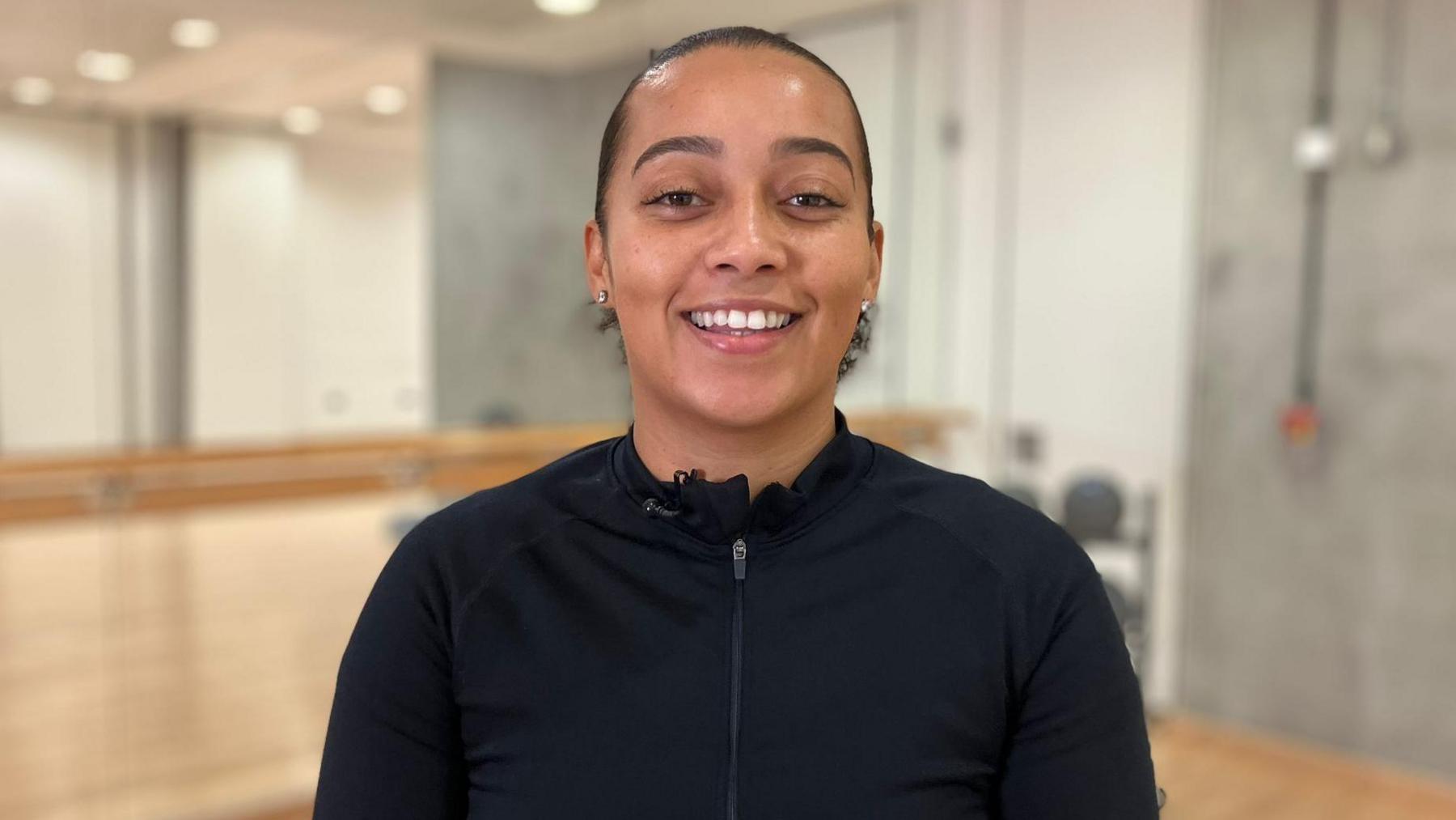 Women's sport advocate Demi Brown stands facing the camera, smiling. She is wearing a zip-up, long-sleeve black sports top.
