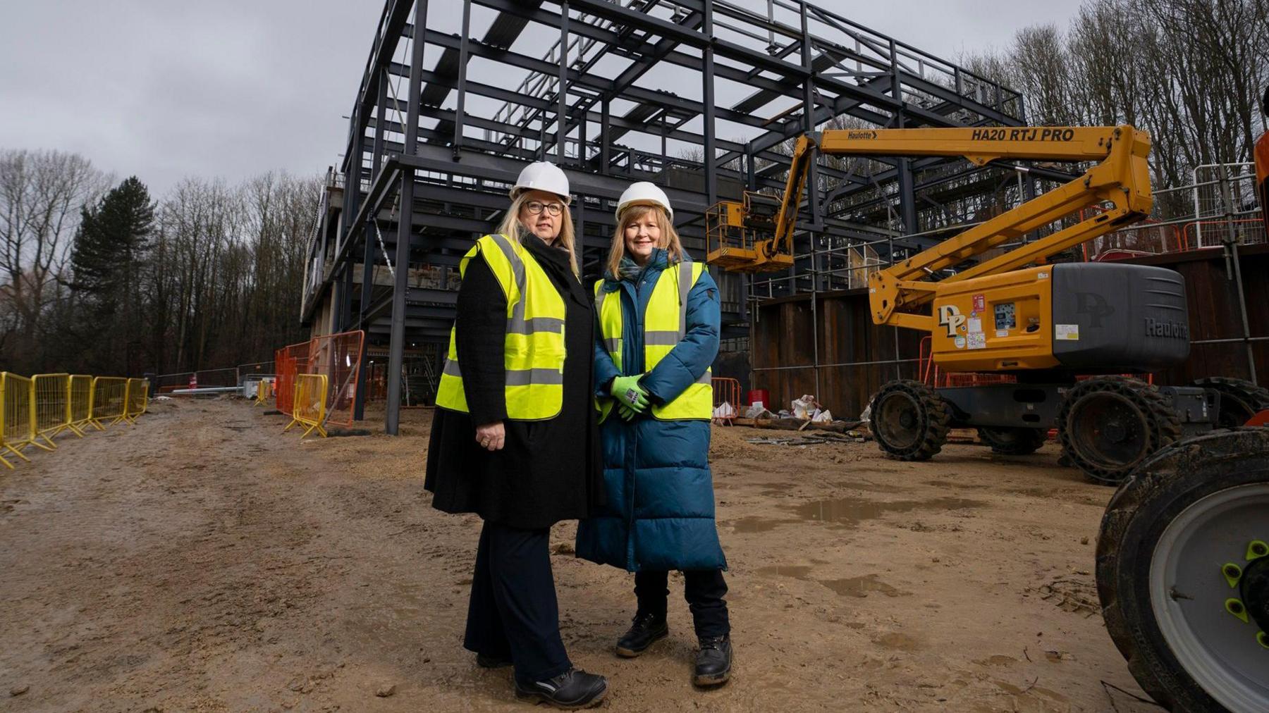 Councillor Elizabeth Scott who has blonde hair and is wearing a white hard hat and yellow hi-vis jacket standing alongside another blonde woman wearing the same safety gear. They're both standing in front of the large, steel frame of the extension. Construction equipment can also be seen.