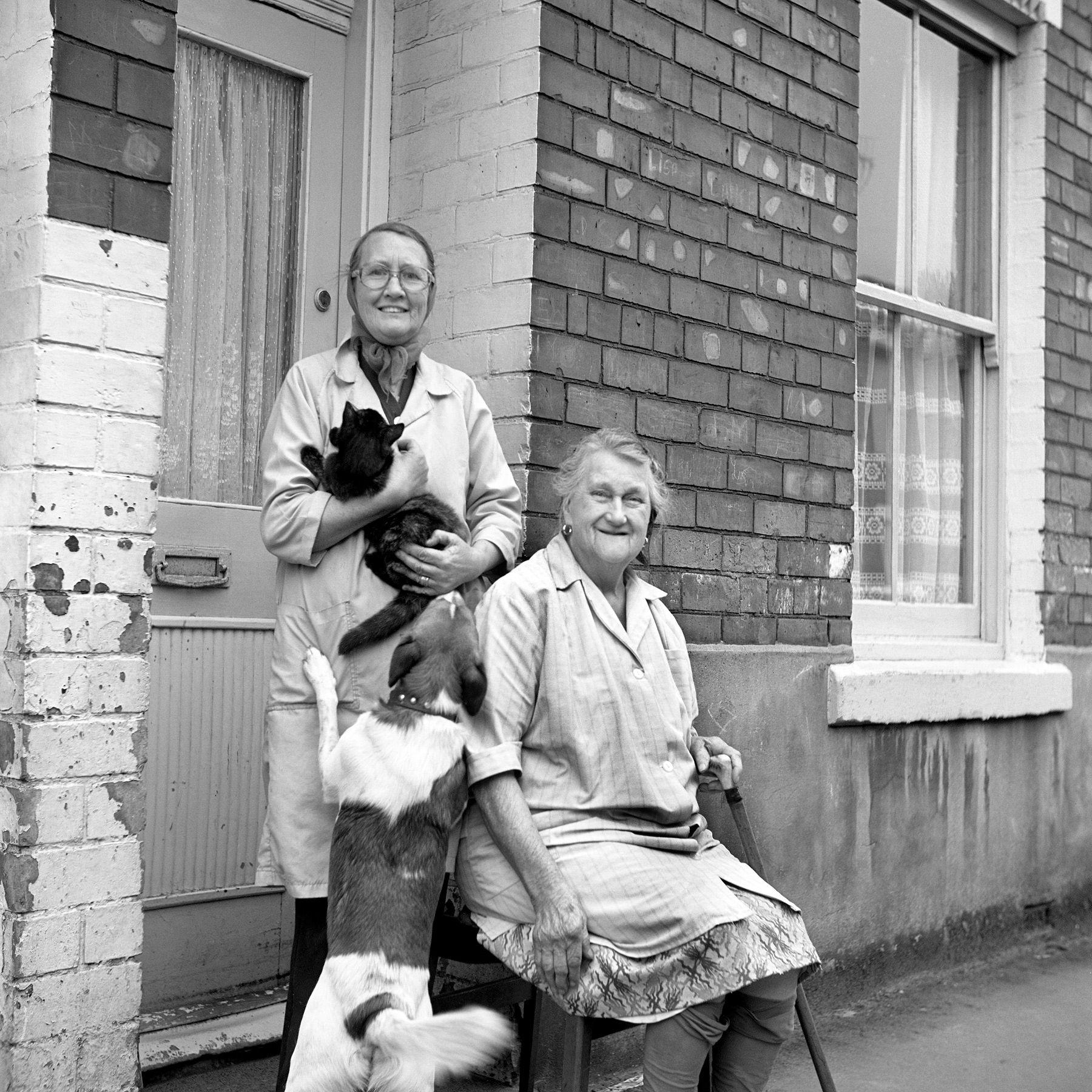 A black and white photograph of two women, one of them elderly, standing outside a brick house. One is holding a cat, smiling, while the other sits with a cane, also smiling. A dog jumps up toward the cat.