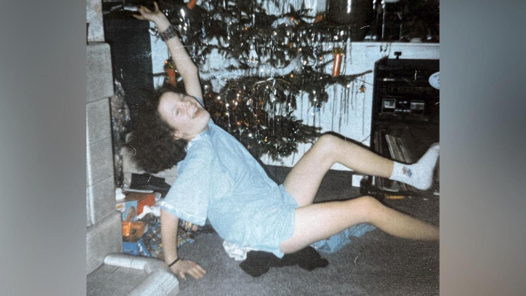 A picture of Lindsay Rimer as a child. She has shoulder length hair and is wearing a blue top and shorts and is sat on the floor with a Christmas tree in the background. 