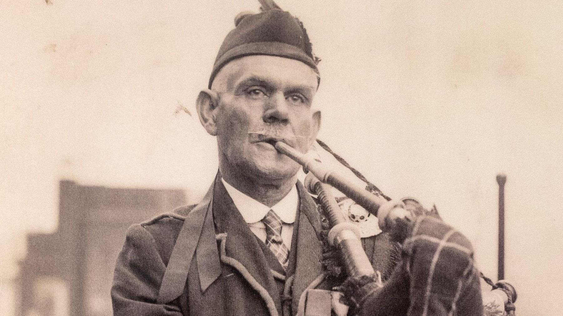 A sepia-hued image of piper Daniel Laidlaw, a smart older man in uniform, playing the bagpipes. He is white-haired, with a Glengarry hat and a white winged moustache.